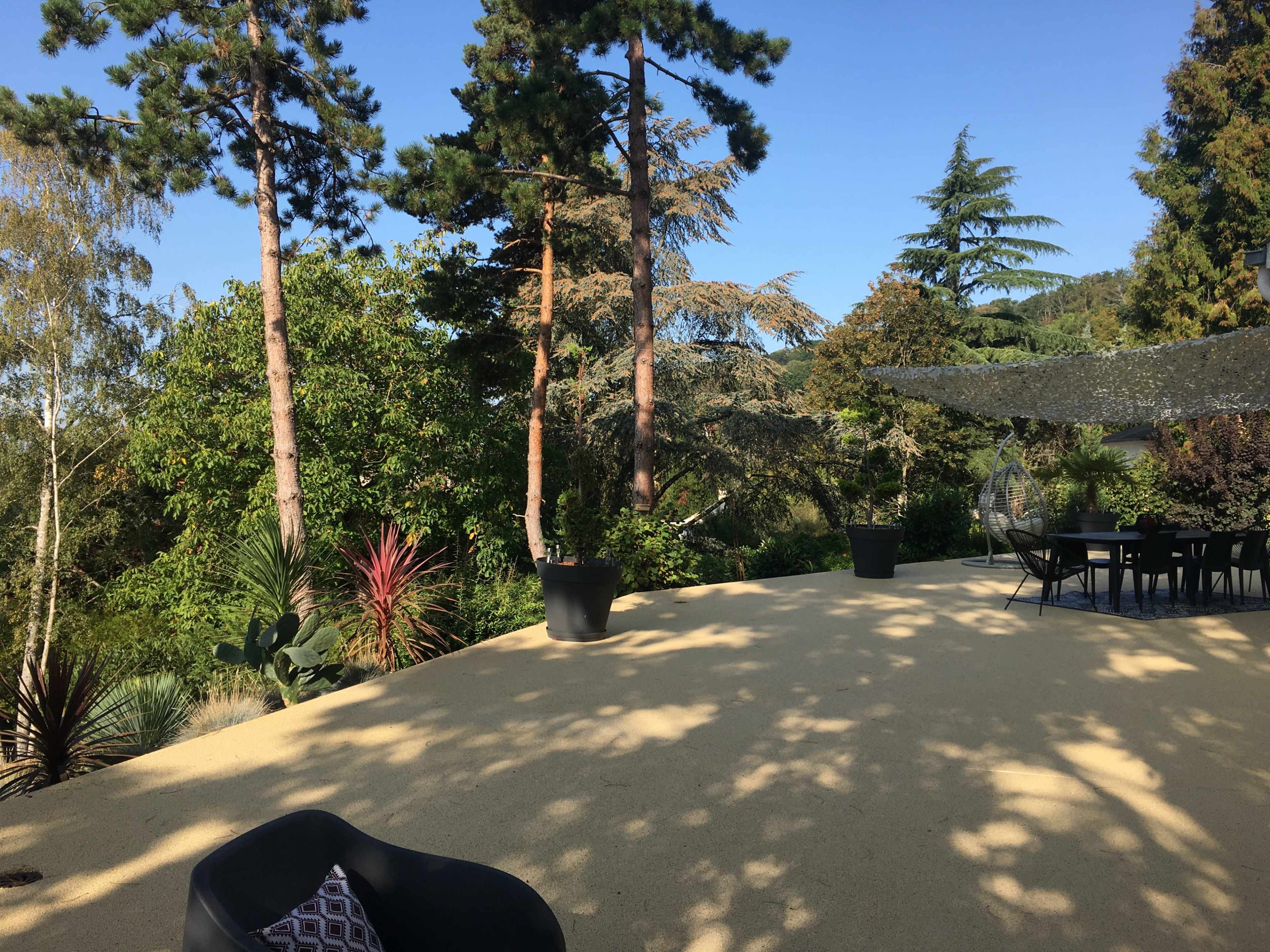 A landscaped outdoor area features trees, plants, and a dining table under a shaded structure.