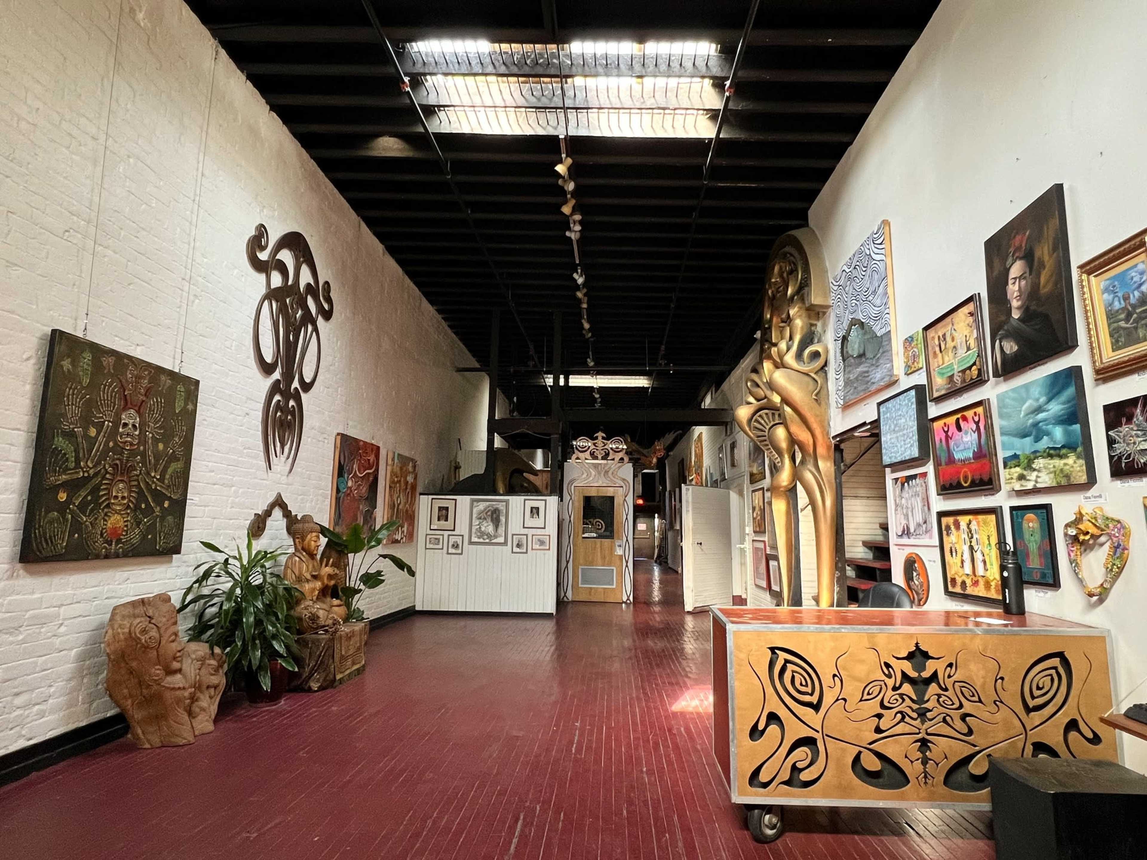 The image shows a spacious art gallery with various artworks displayed on the walls and a wooden reception desk in the foreground.