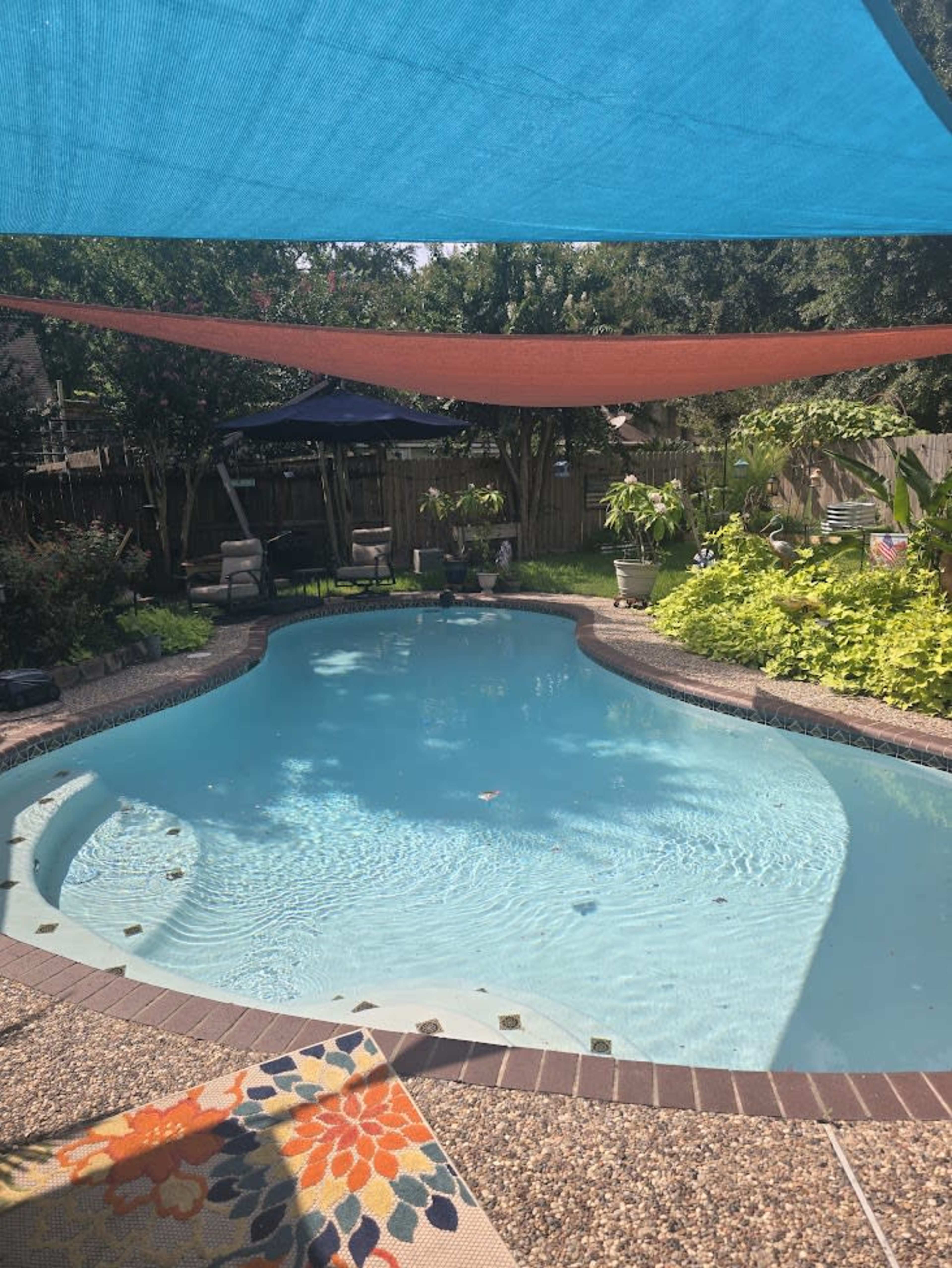 A backyard pool is shaded by colorful awnings, surrounded by greenery and outdoor furniture.