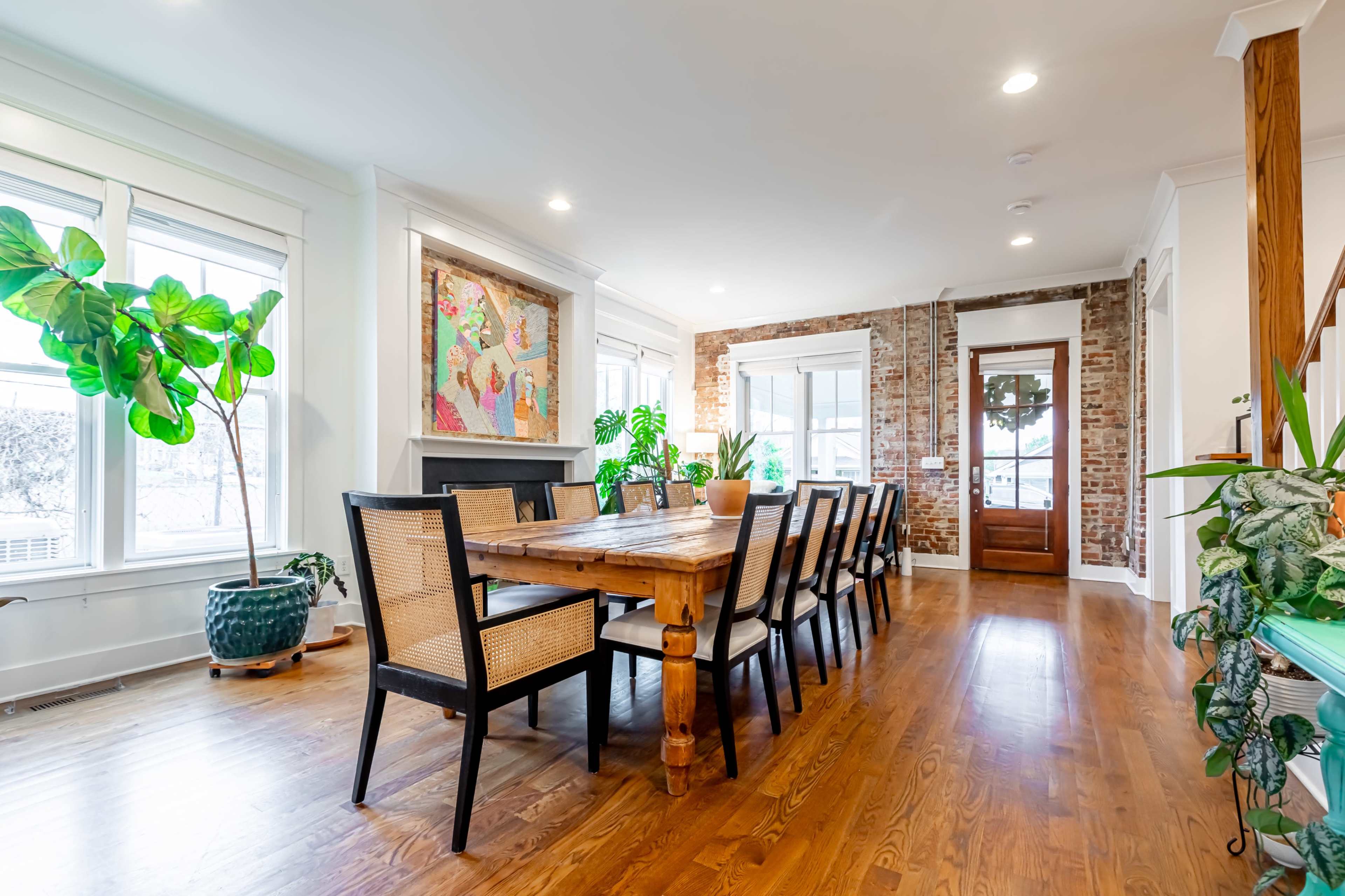 The spacious dining area features a long wooden table surrounded by black chairs, with large windows providing natural light, and a mix of plants and artwork adorning the walls.