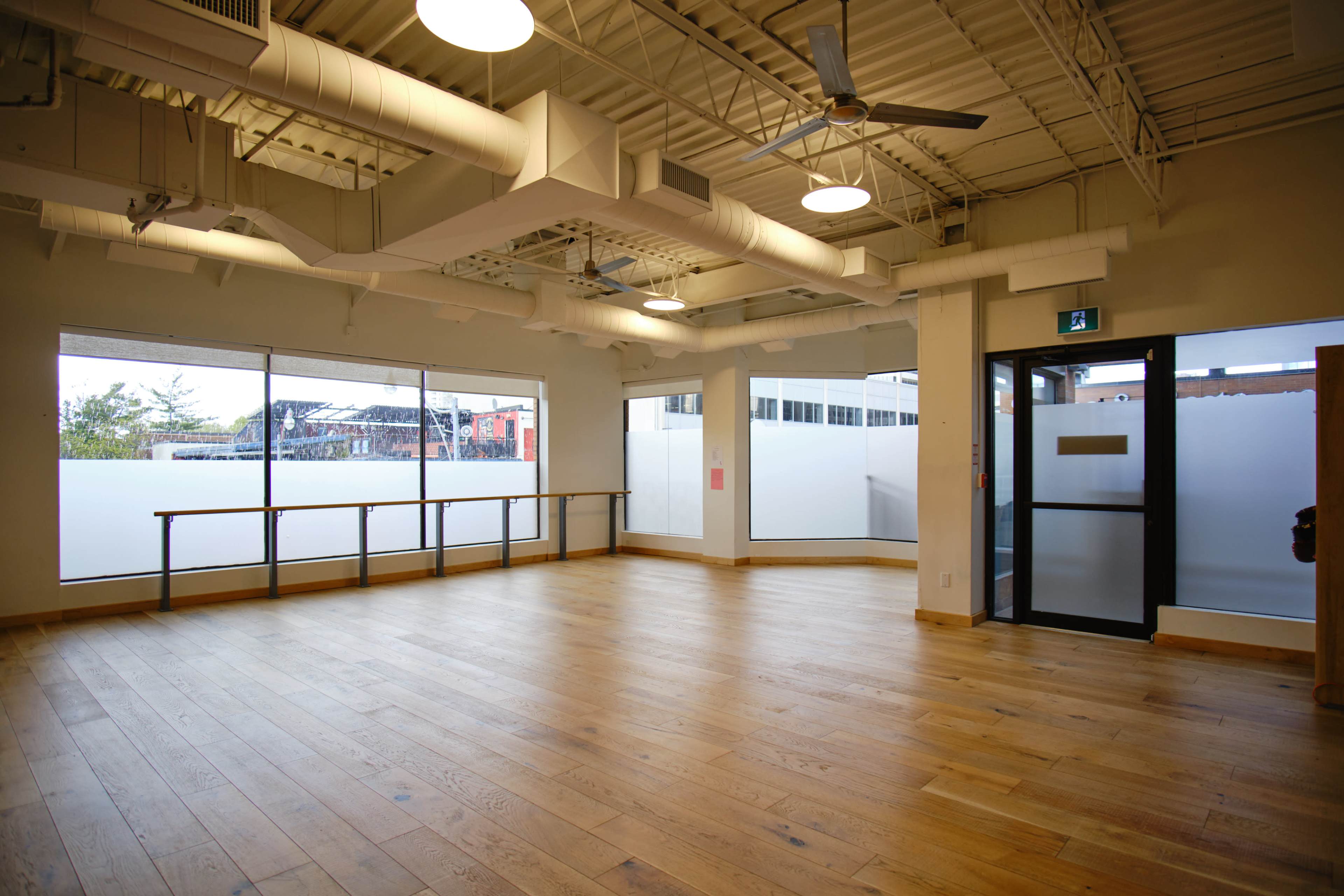 The image shows an empty room with wooden flooring, large windows, and a door leading outside.