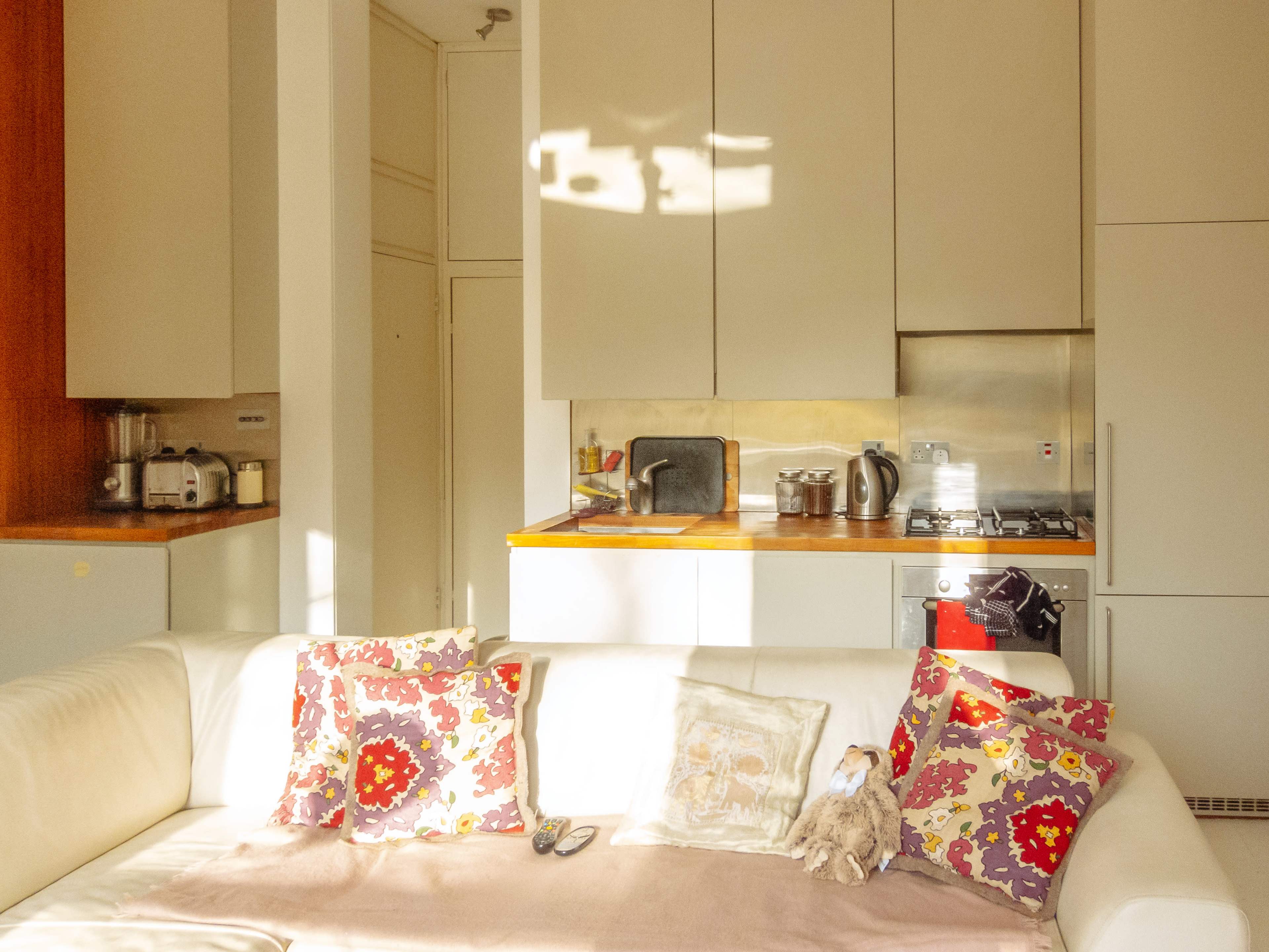 A light-filled kitchen and living area feature a white couch adorned with colorful pillows and a view of appliances and cabinetry in the background.