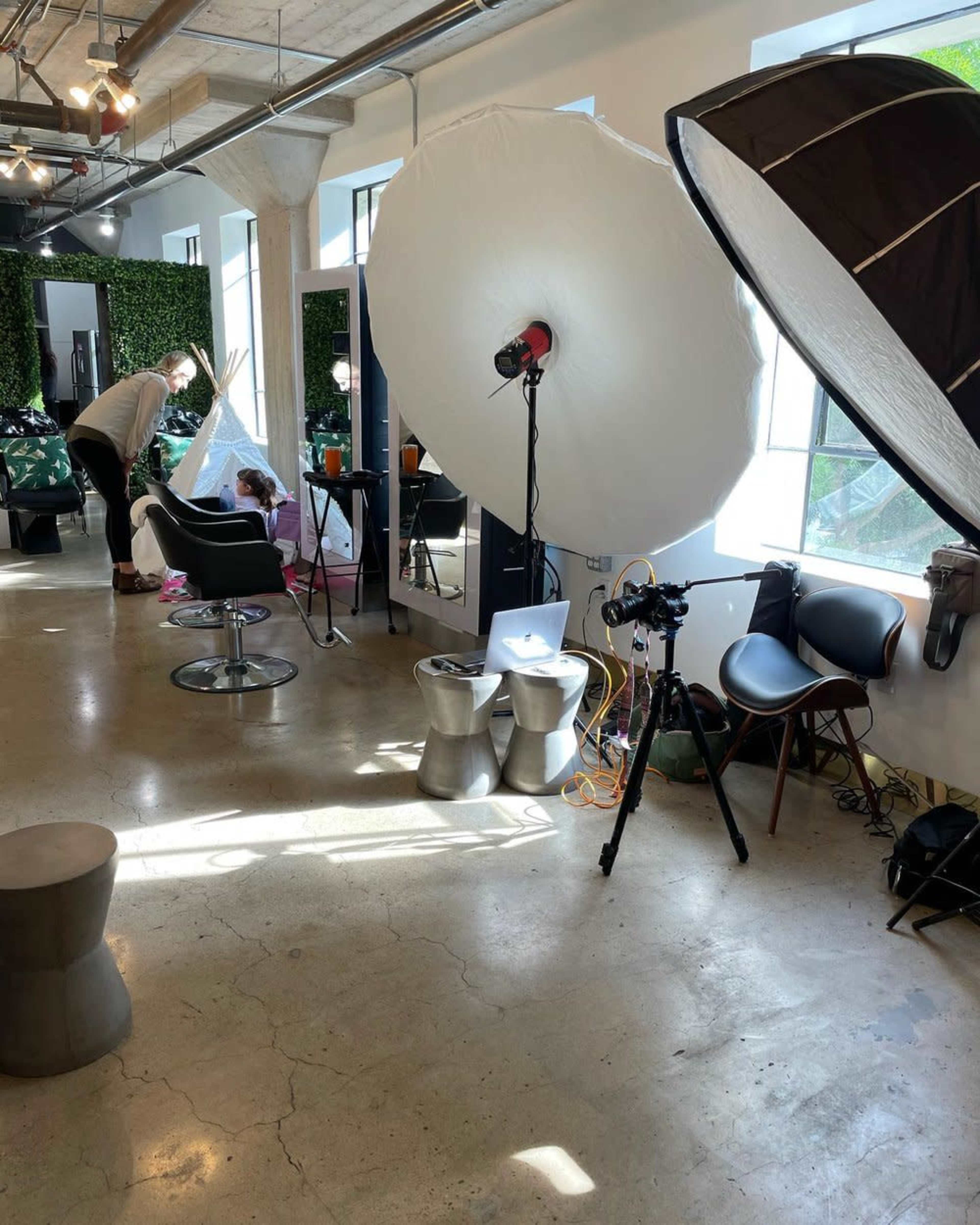 A photography studio is set up with a large softbox light, a camera on a tripod, and a person arranging props while another individual sits in a styling chair.