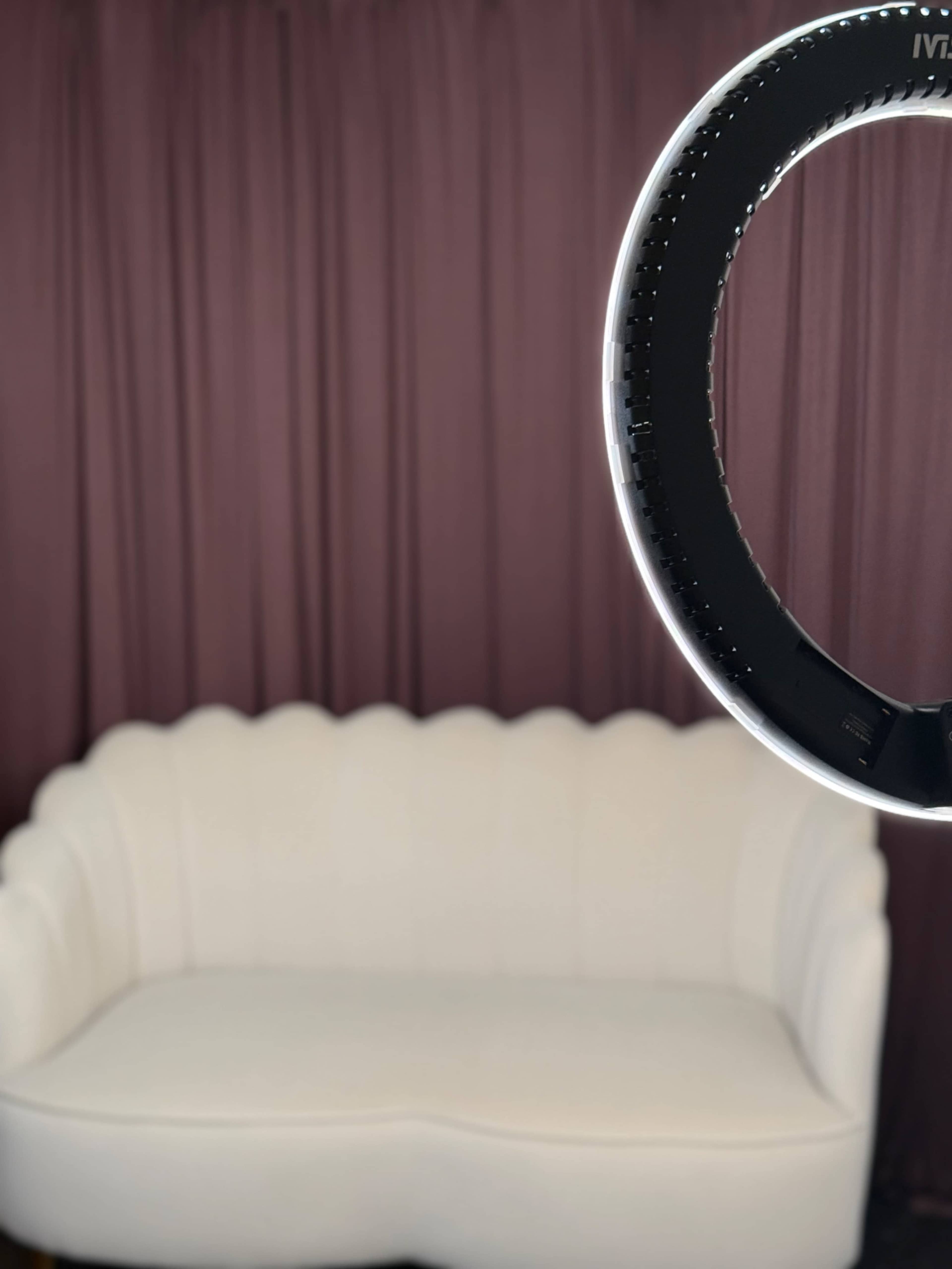 A white couch is positioned in front of a purple curtain, with a ring light partially visible in the foreground.