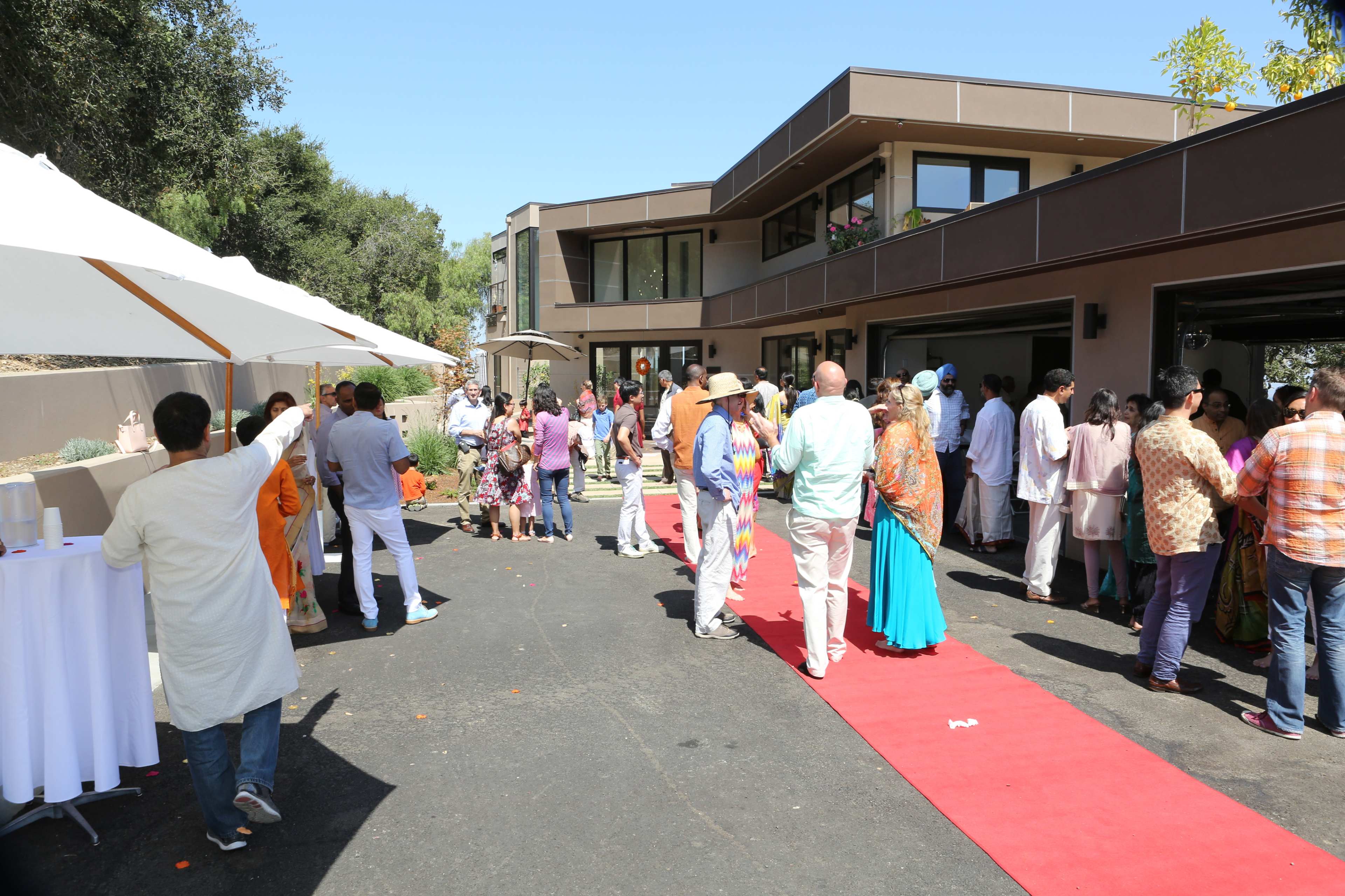 A group of people in traditional attire gather at an outdoor event near a modern building, with a red carpet and white umbrellas set up.