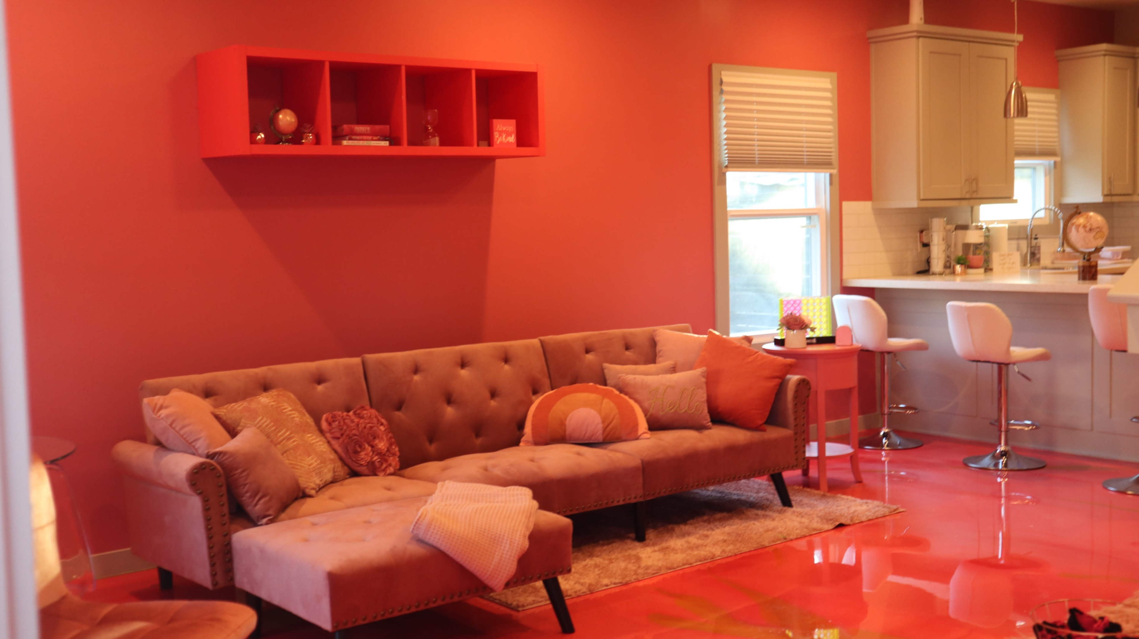 A living room with a pink wall, a gray tufted sofa, and a red shelf above it, accompanied by a small round table and bar stools in the background.