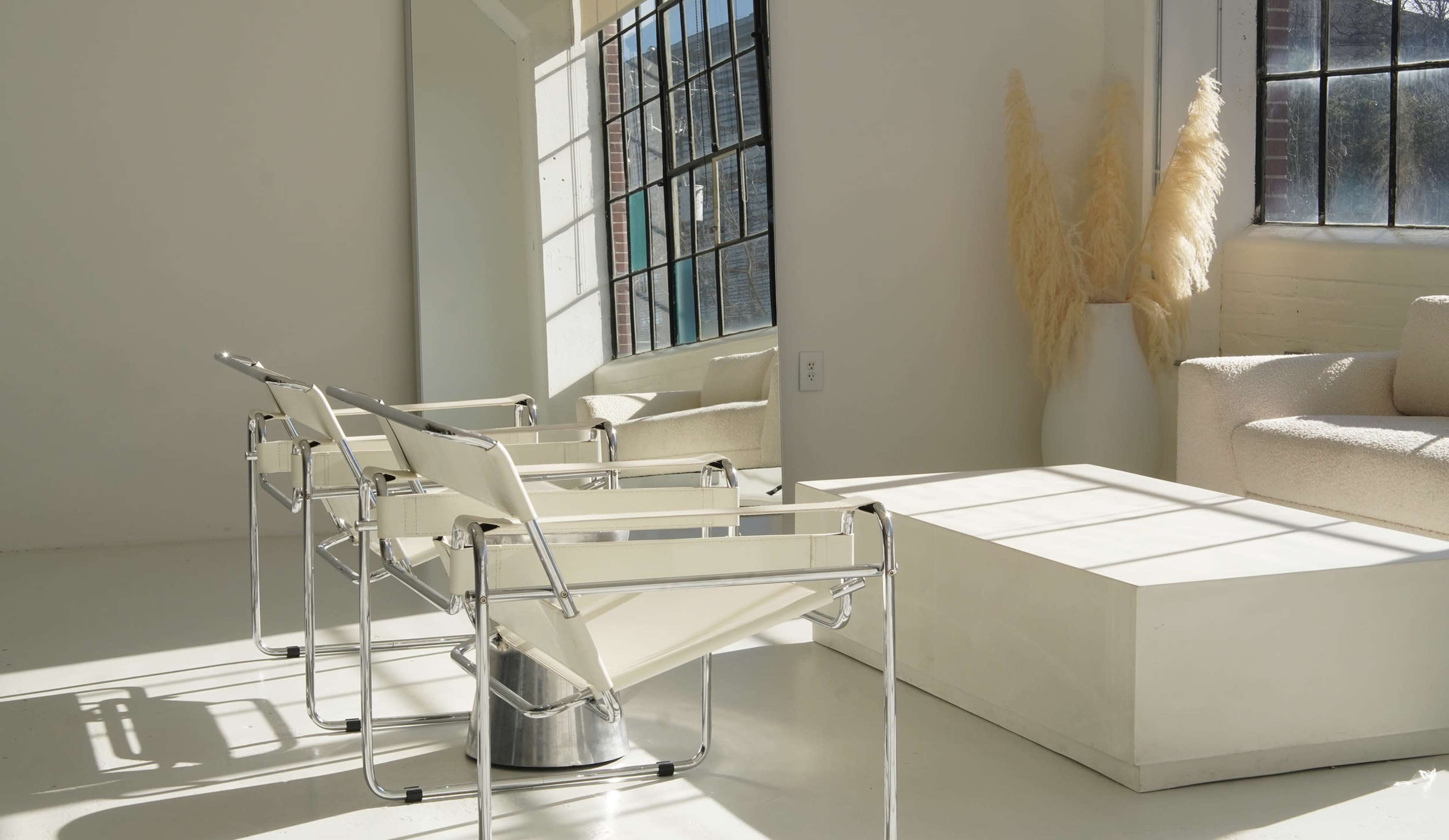 Two modern chairs facing a minimalist coffee table, with a large mirror reflecting light from a window in a bright interior space.