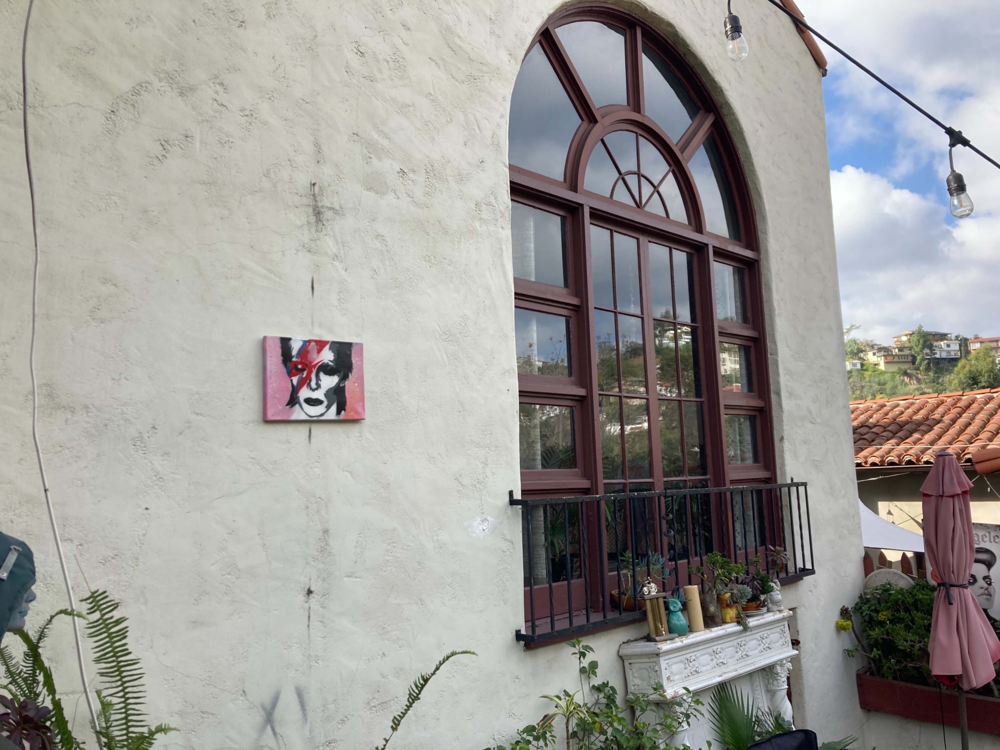 A wall with a large arched window features a painting of a woman and decorative plants below it.