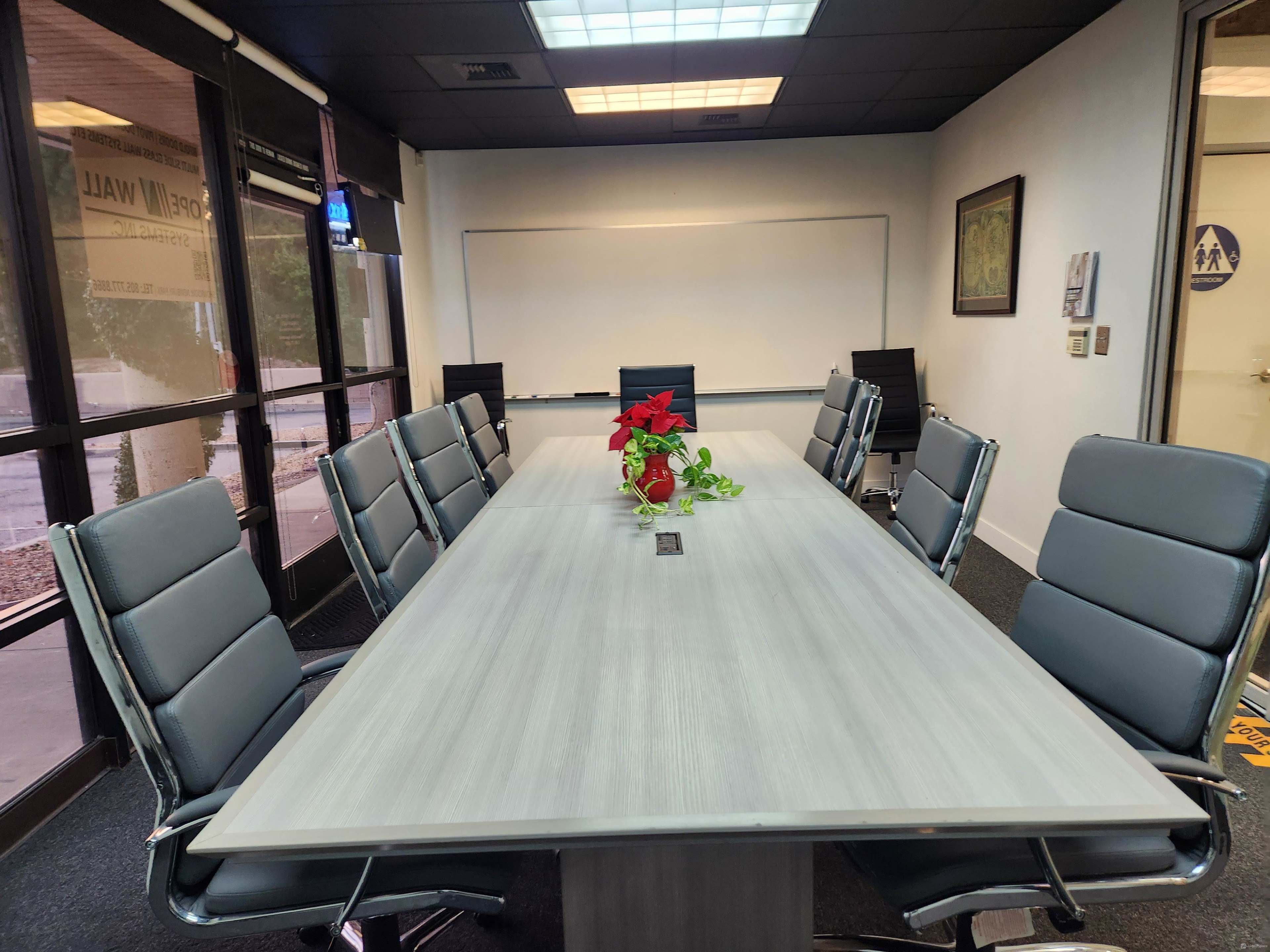 The image shows a conference room with a large rectangular table surrounded by eight gray chairs, and a red plant centerpiece on the table.
