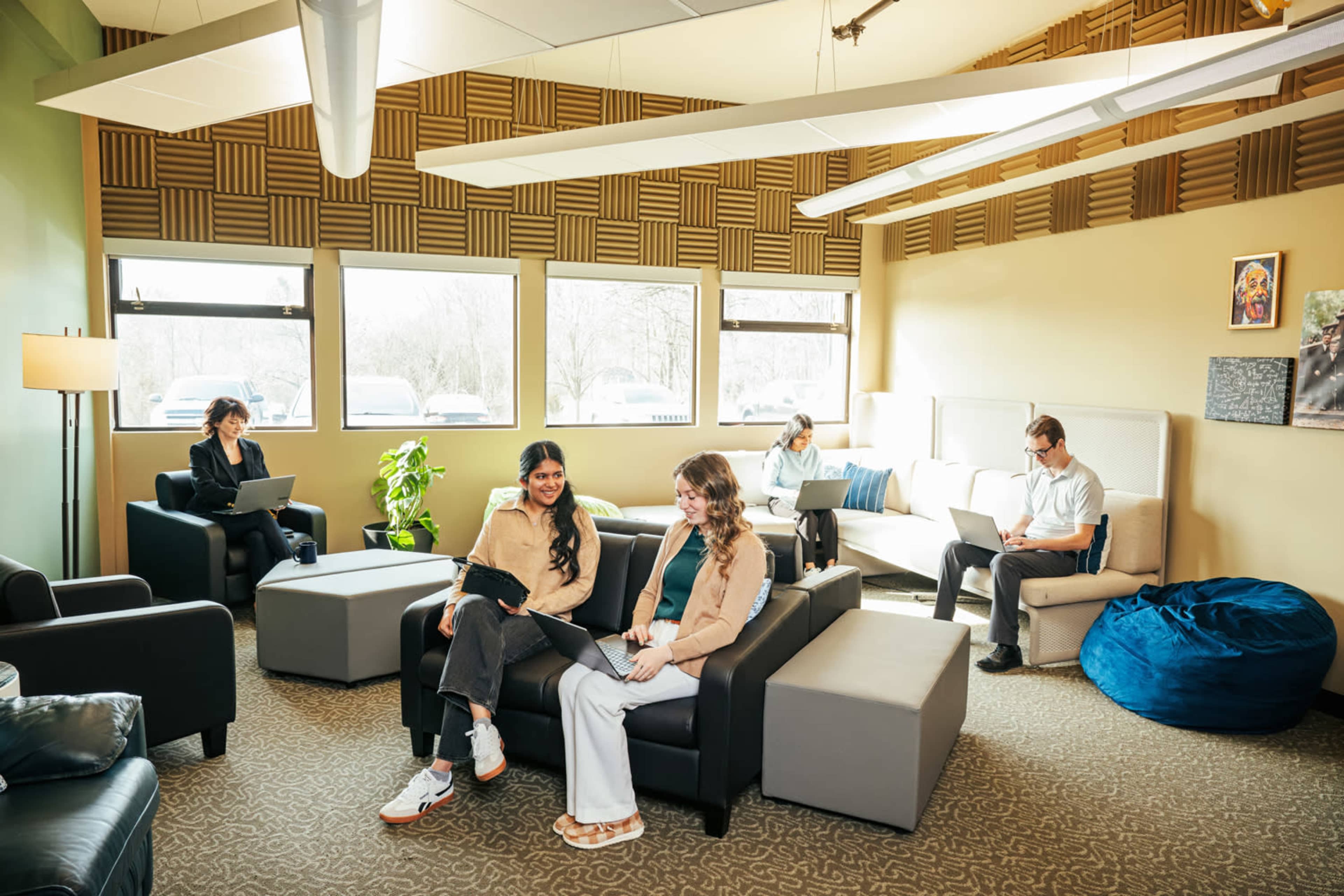 A group of five people are working or relaxing in a modern lounge area furnished with sofas, chairs, and a beanbag.