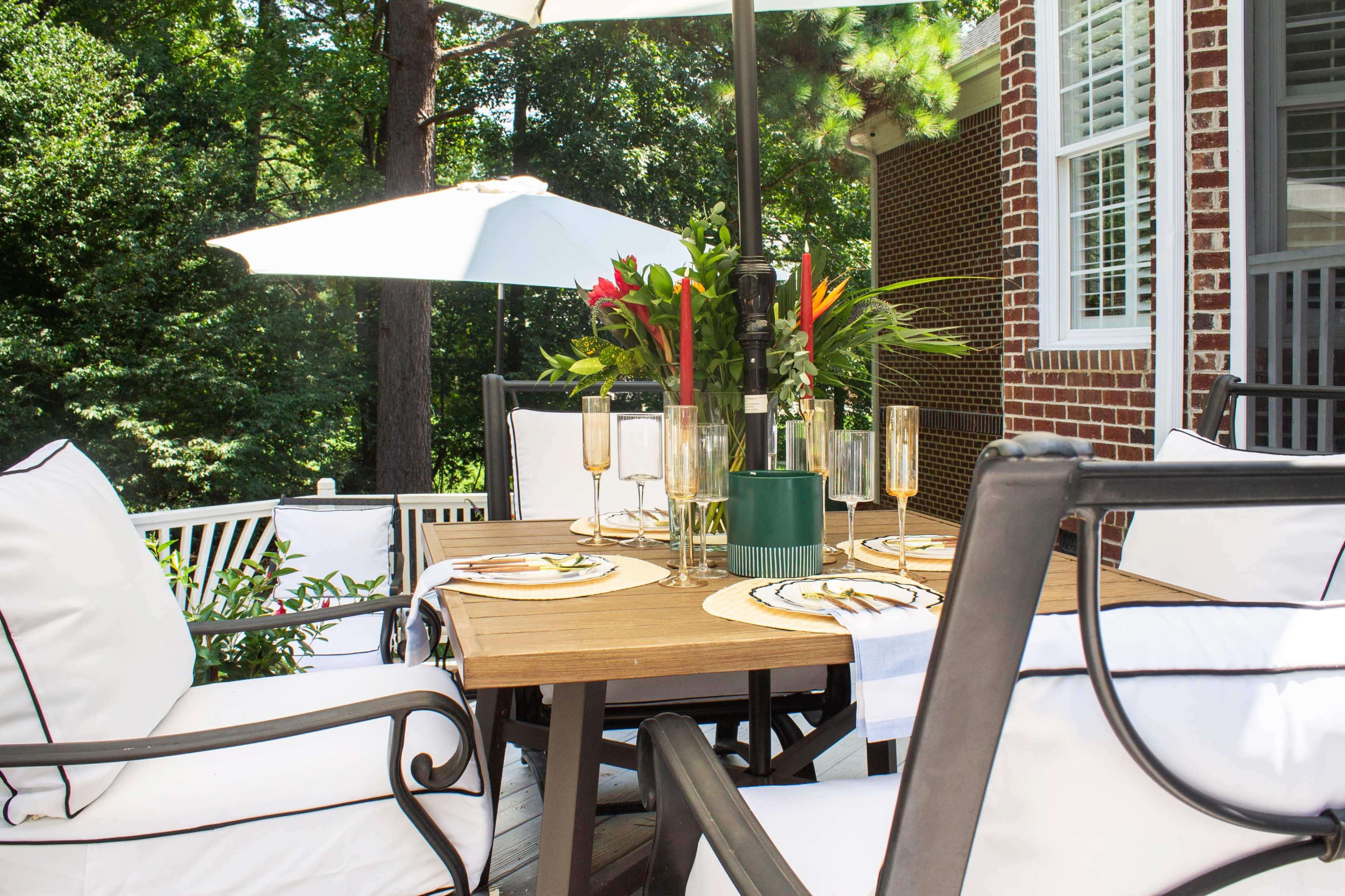 A patio dining area features a wooden table surrounded by cushioned chairs, with an umbrella overhead and tropical plants in the background.