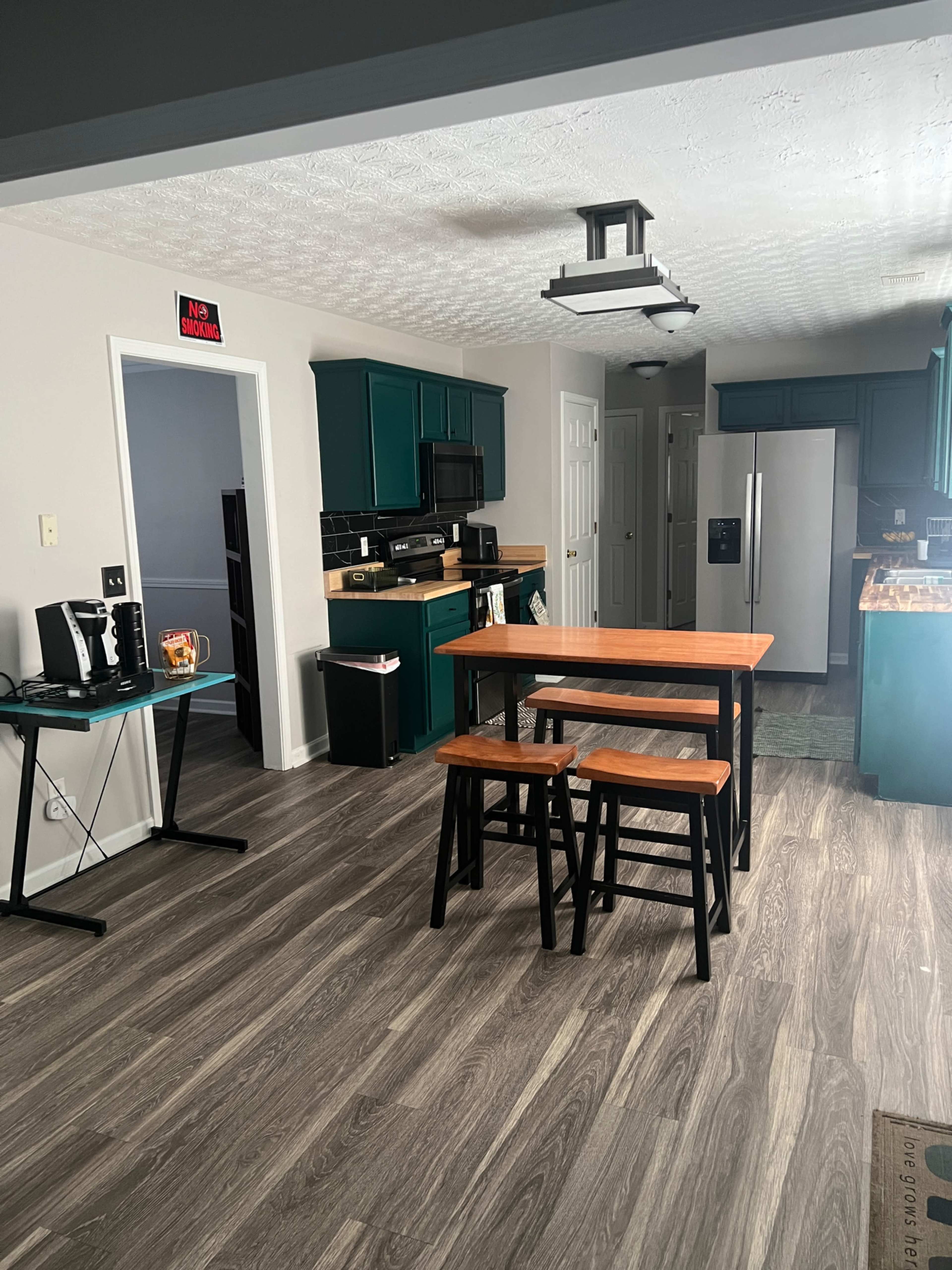 The image shows a modern kitchen with teal cabinets, a stainless steel refrigerator, and a wooden table with black stools in the center.