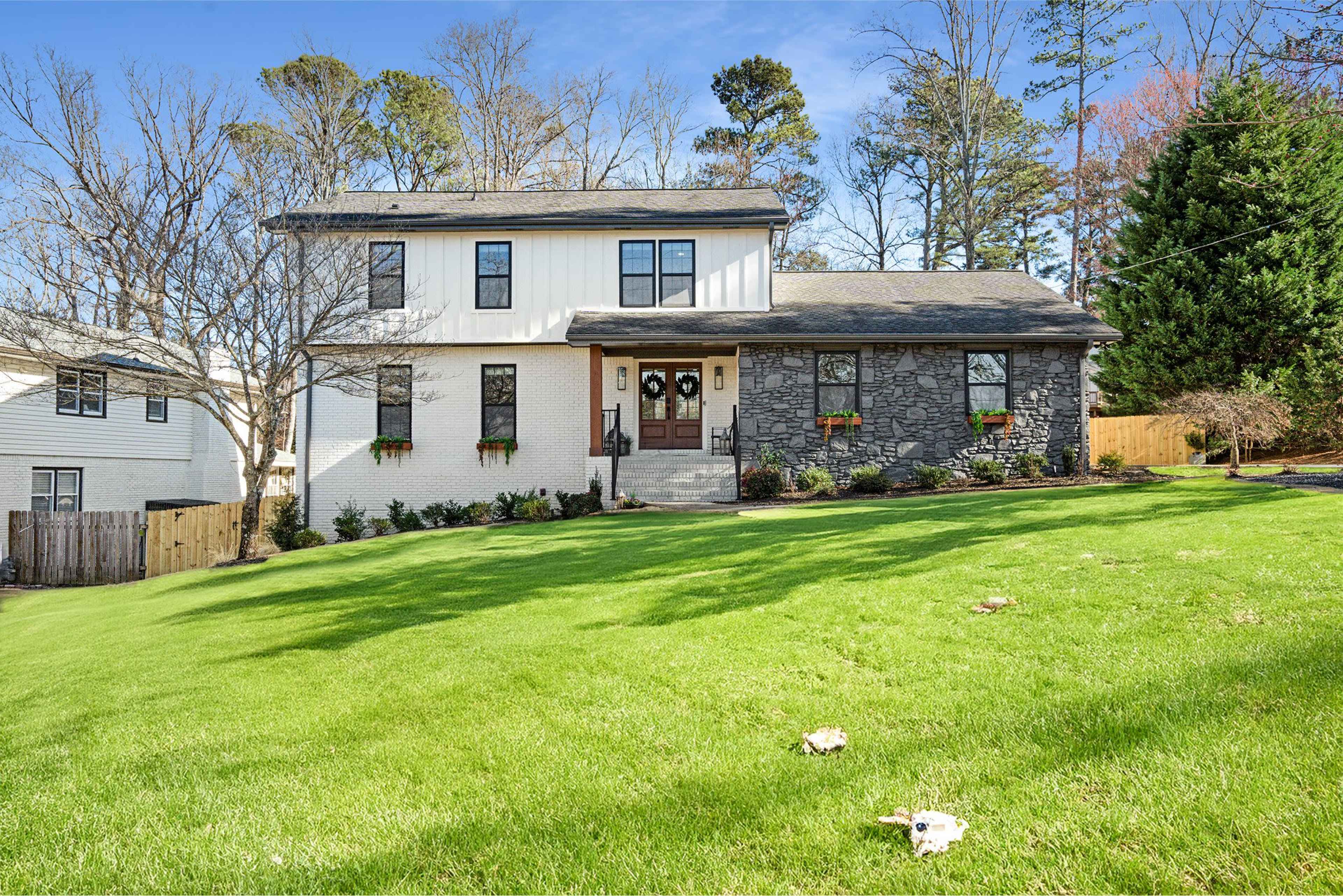 The image shows a two-story house featuring a combination of white exterior siding and stone accents, situated on a grassy lawn with trees in the background.