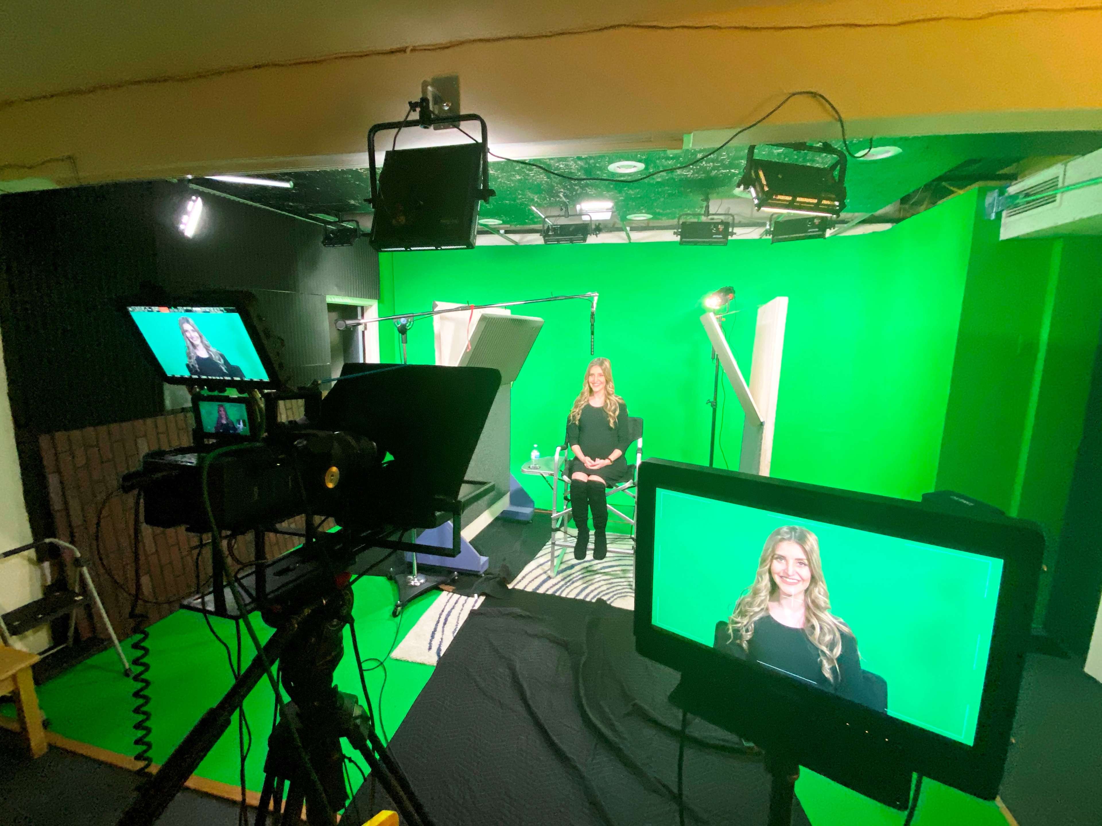 A woman with long hair sits in front of a green screen in a video production studio, surrounded by multiple cameras and lights.