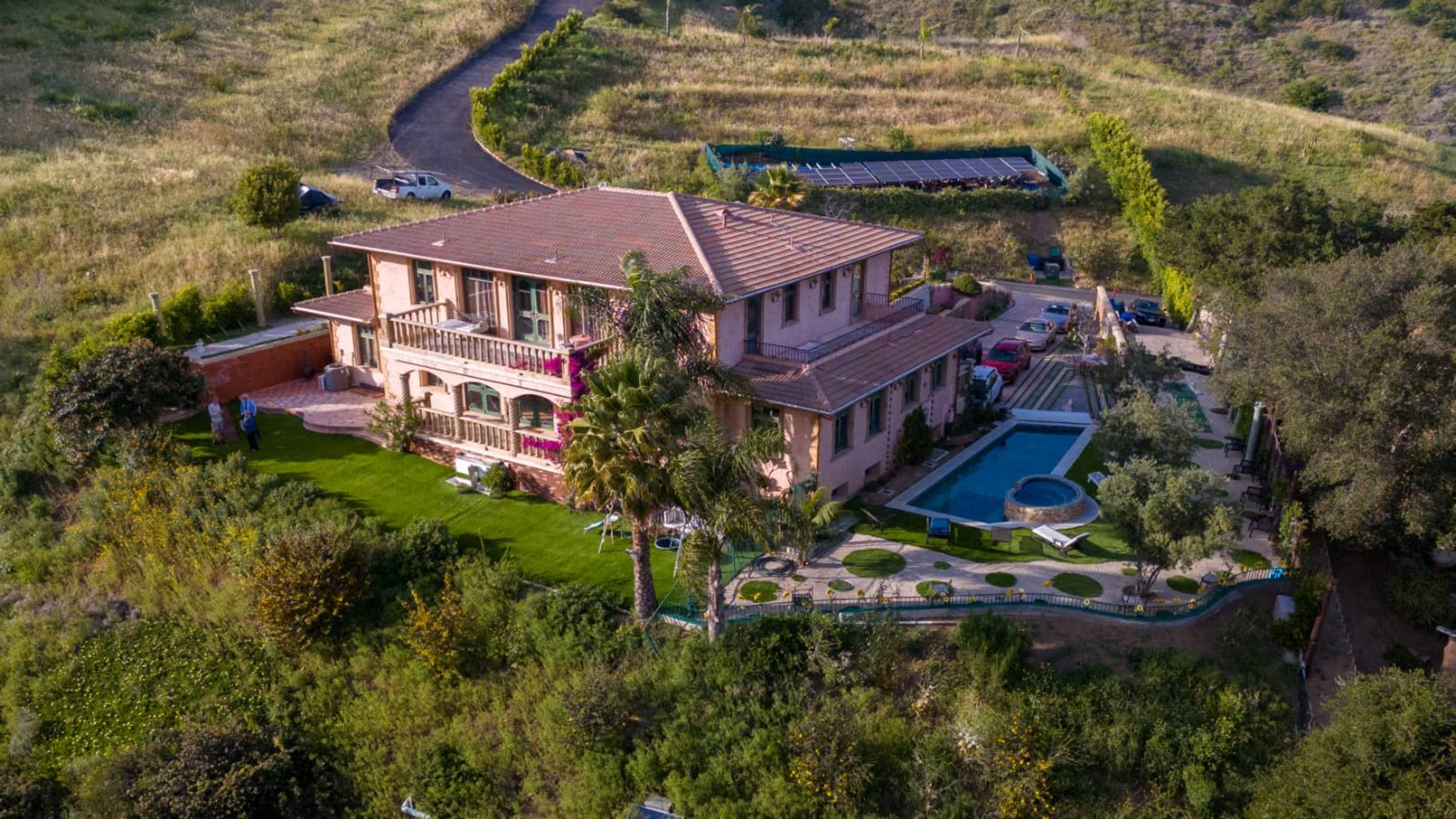 The image shows a large two-story house with a pool and landscaped gardens, surrounded by greenery and a driveway.