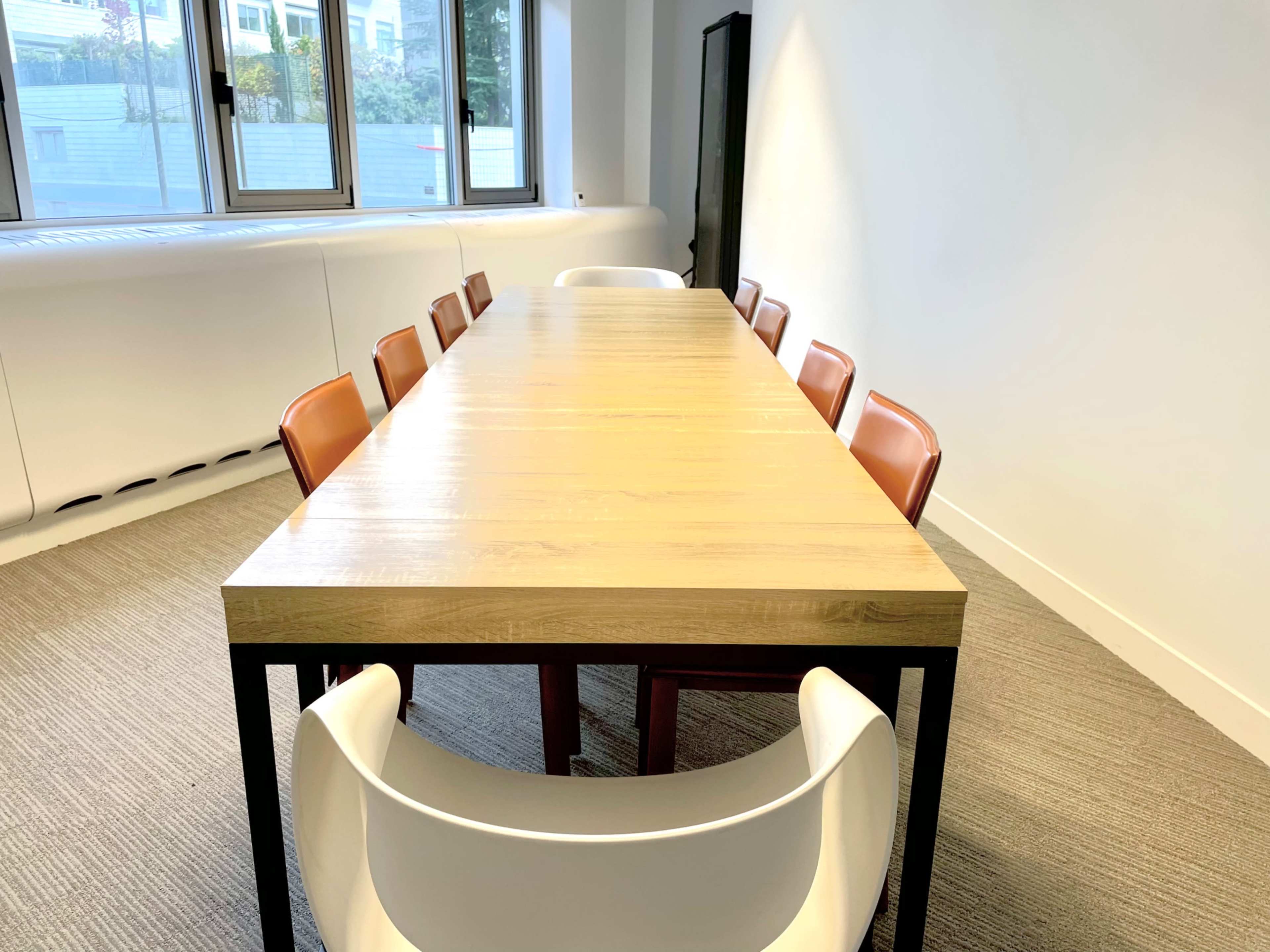 A wooden conference table is surrounded by eight chairs in a well-lit room with large windows.