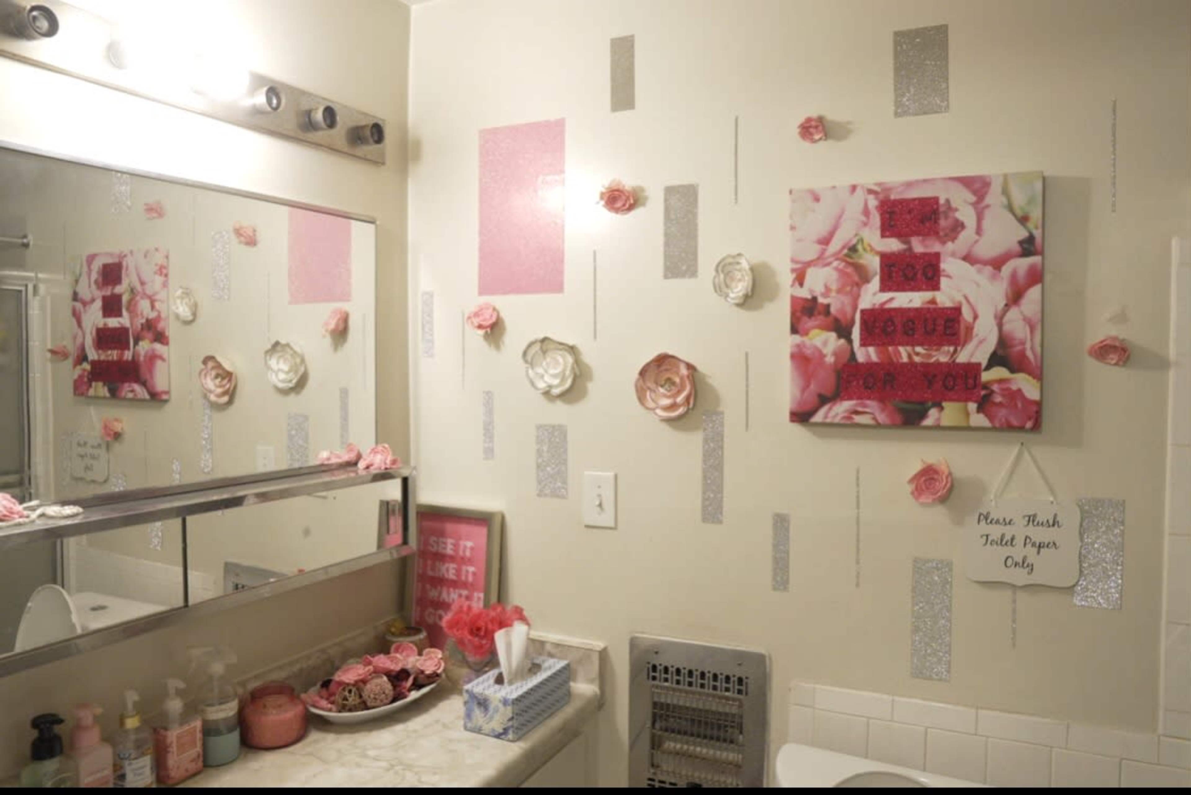 A bathroom features a decorative arrangement of pink roses and glittery accents on the walls, with a framed artwork displayed above a vanity.