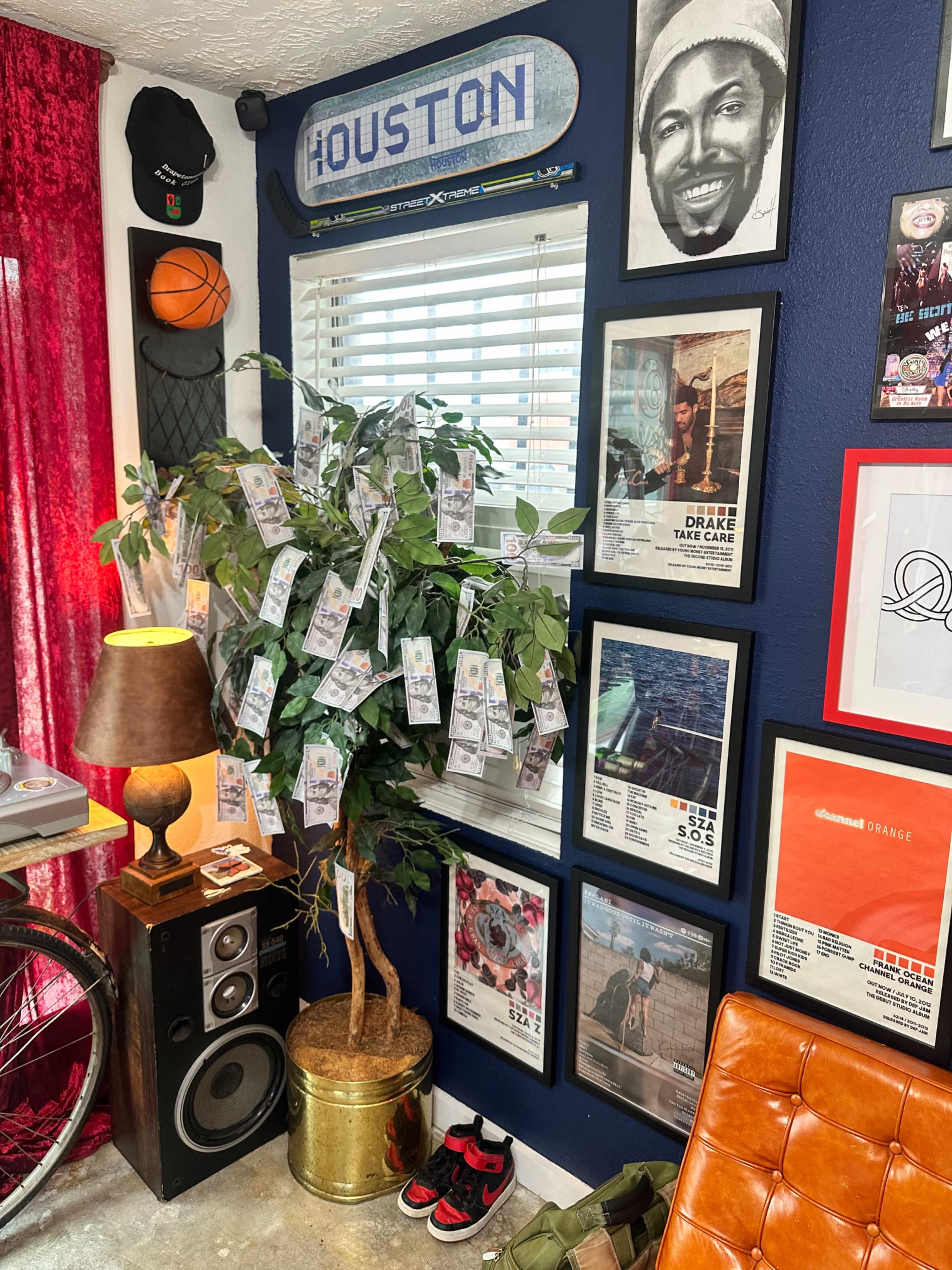 The image shows a corner of a room featuring a small artificial tree adorned with money-themed decorations, surrounded by framed artwork, a lamp, a speaker, and sports memorabilia.