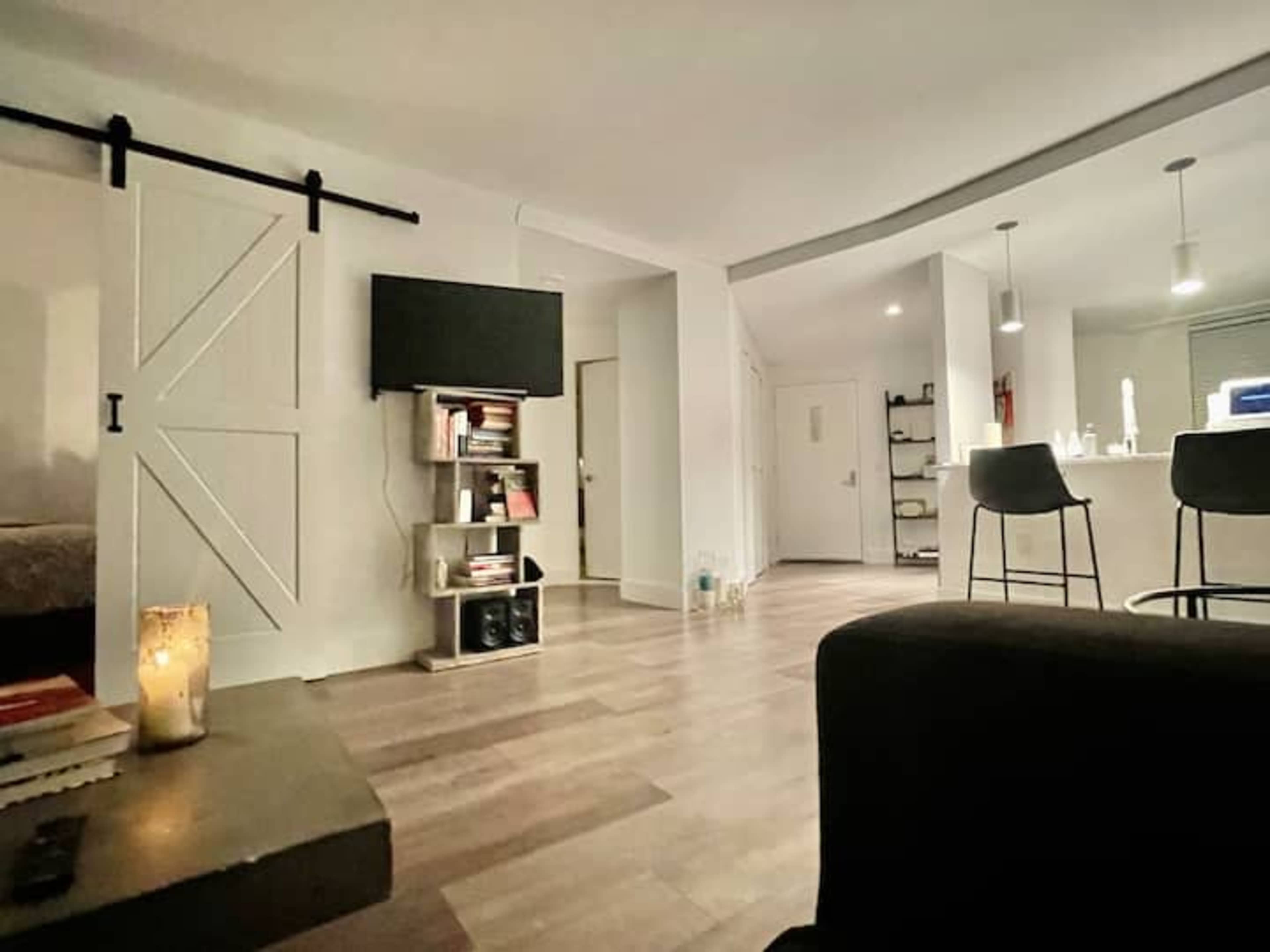 A modern living space features a dark sofa, a bookshelf with books, a TV mounted on the wall, and a kitchen area with bar stools and pendant lighting.