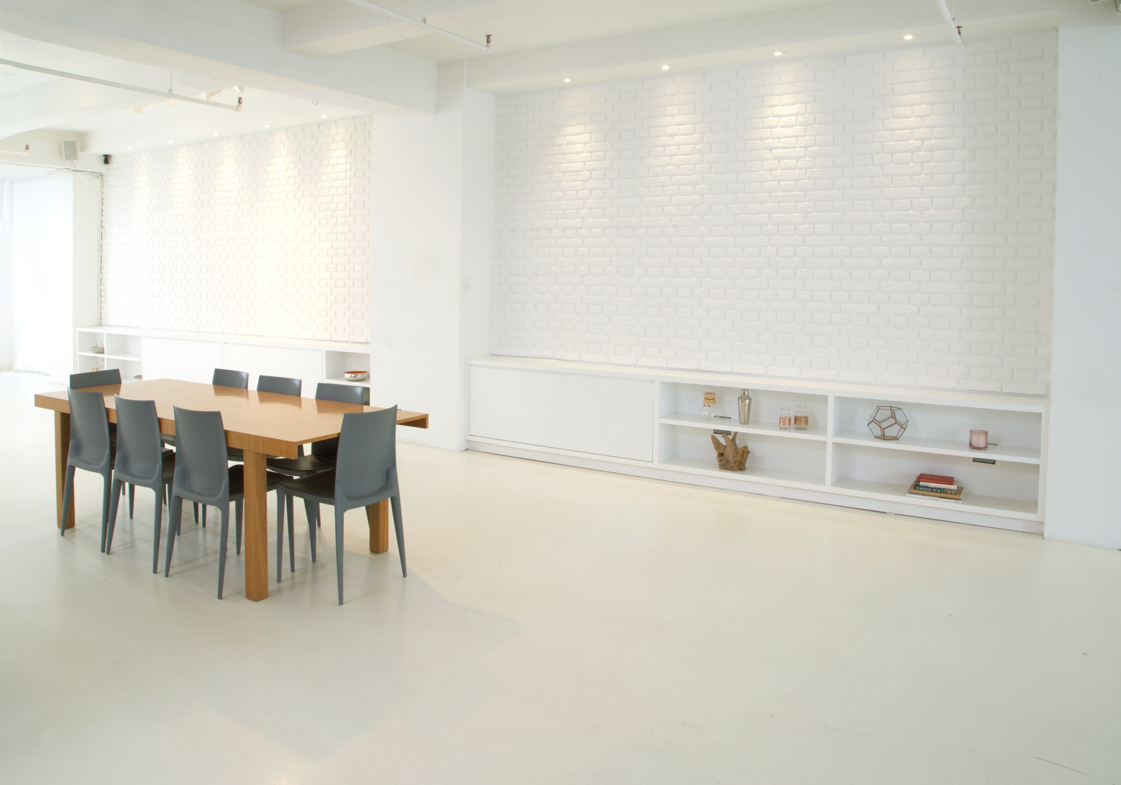 A large wooden dining table with gray chairs is set in a minimalist room featuring a white brick wall and a low shelf displaying decorative items.