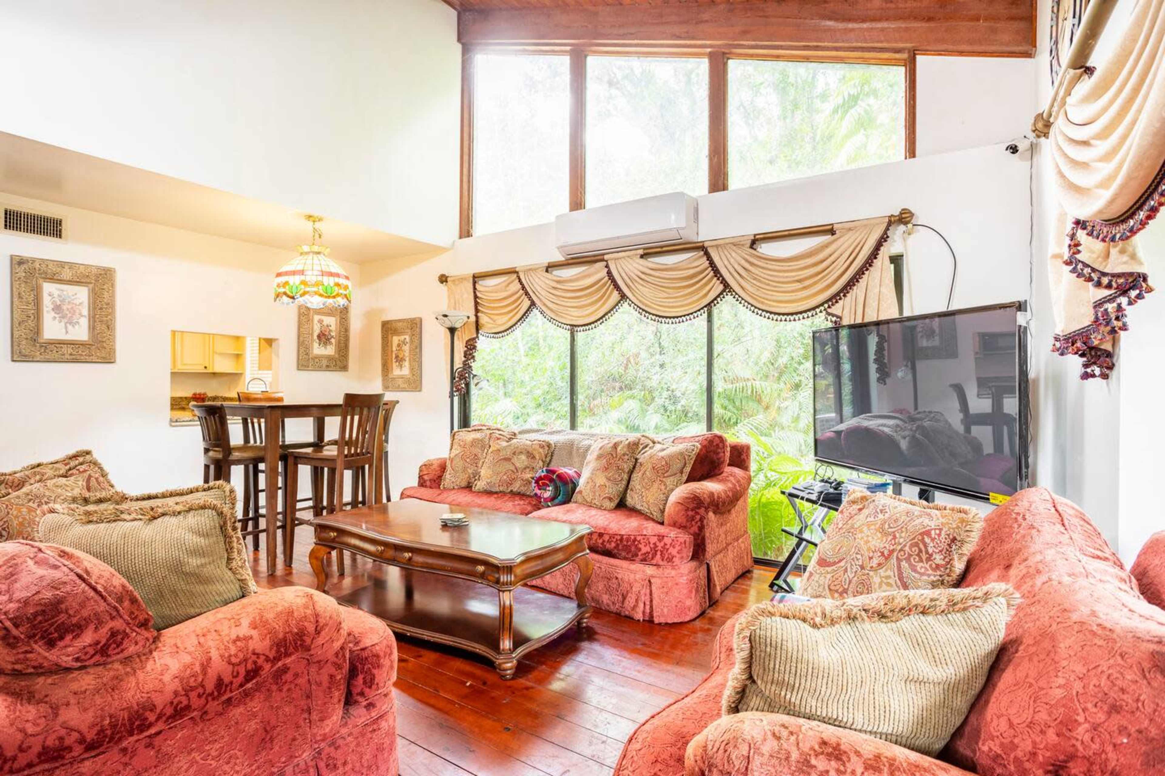 The image shows a living room with red upholstered sofas, a wooden coffee table, and large windows overlooking greenery.