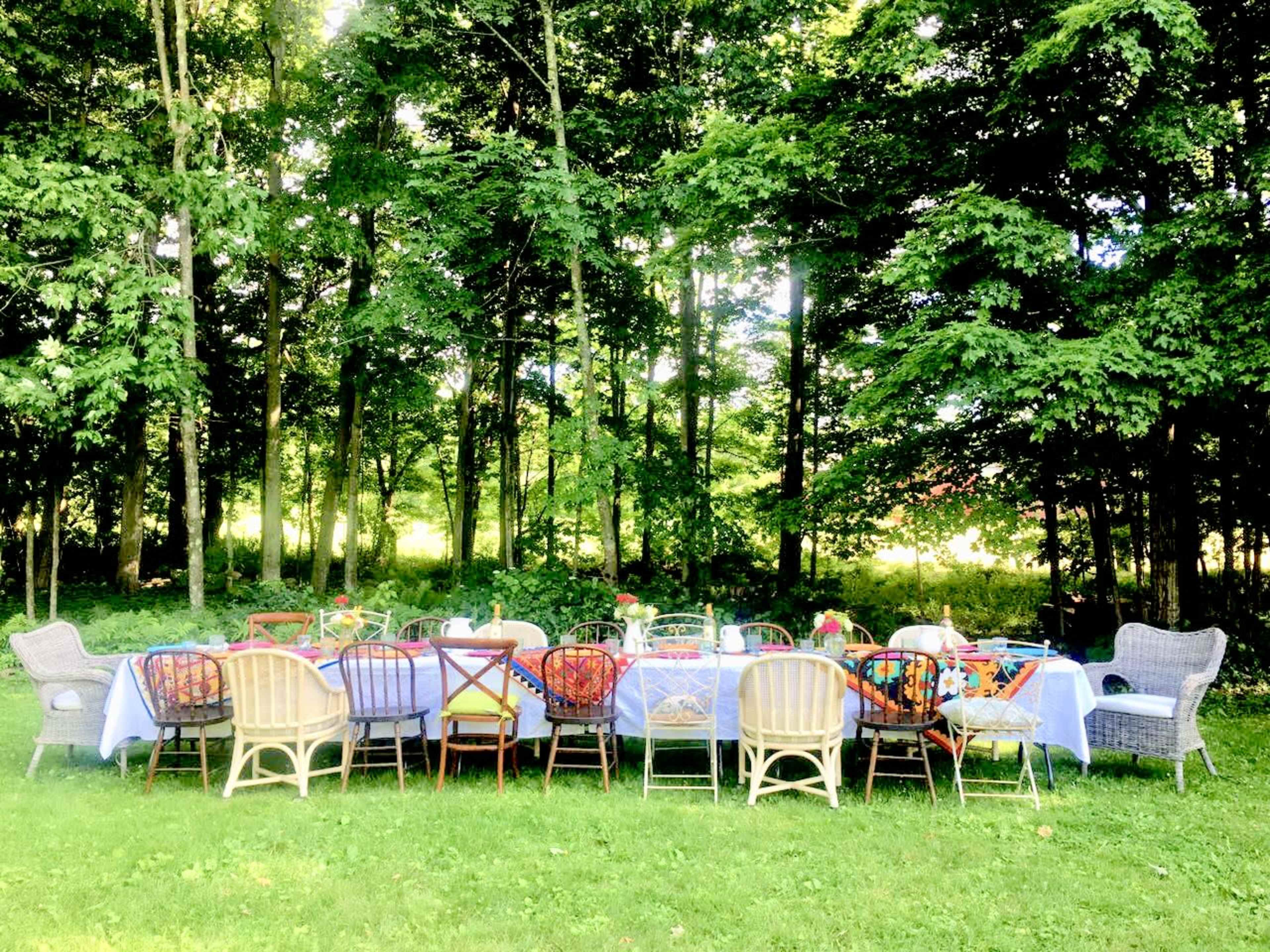 A long table set for a meal is surrounded by various chairs in a wooded outdoor setting.