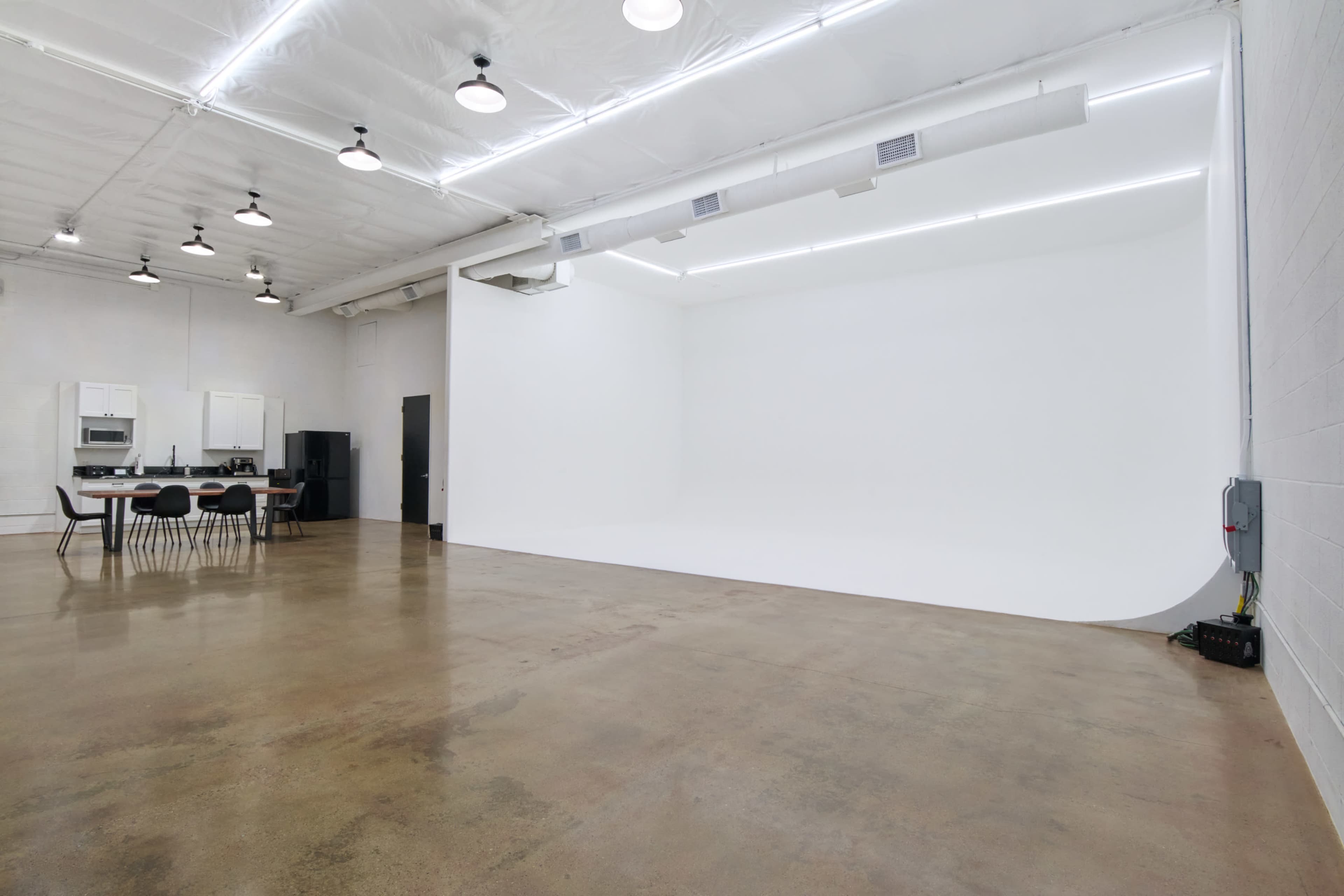 The image shows a spacious studio with a white backdrop, polished concrete floor, and a kitchenette area with black chairs and cabinets in the corner.