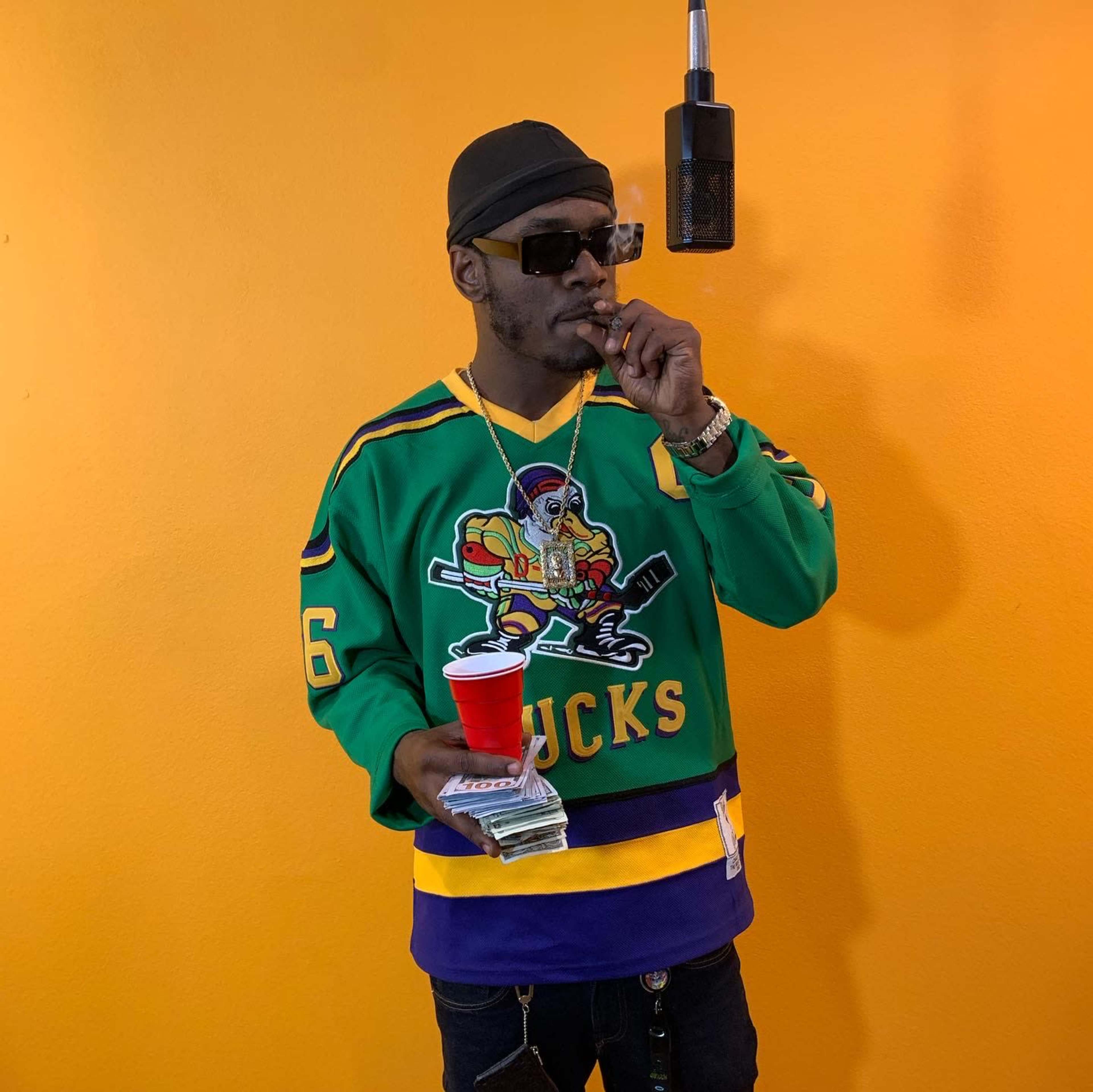 A person wearing a colorful hockey jersey holds a red cup and a stack of cash while smoking a cigar against an orange wall.