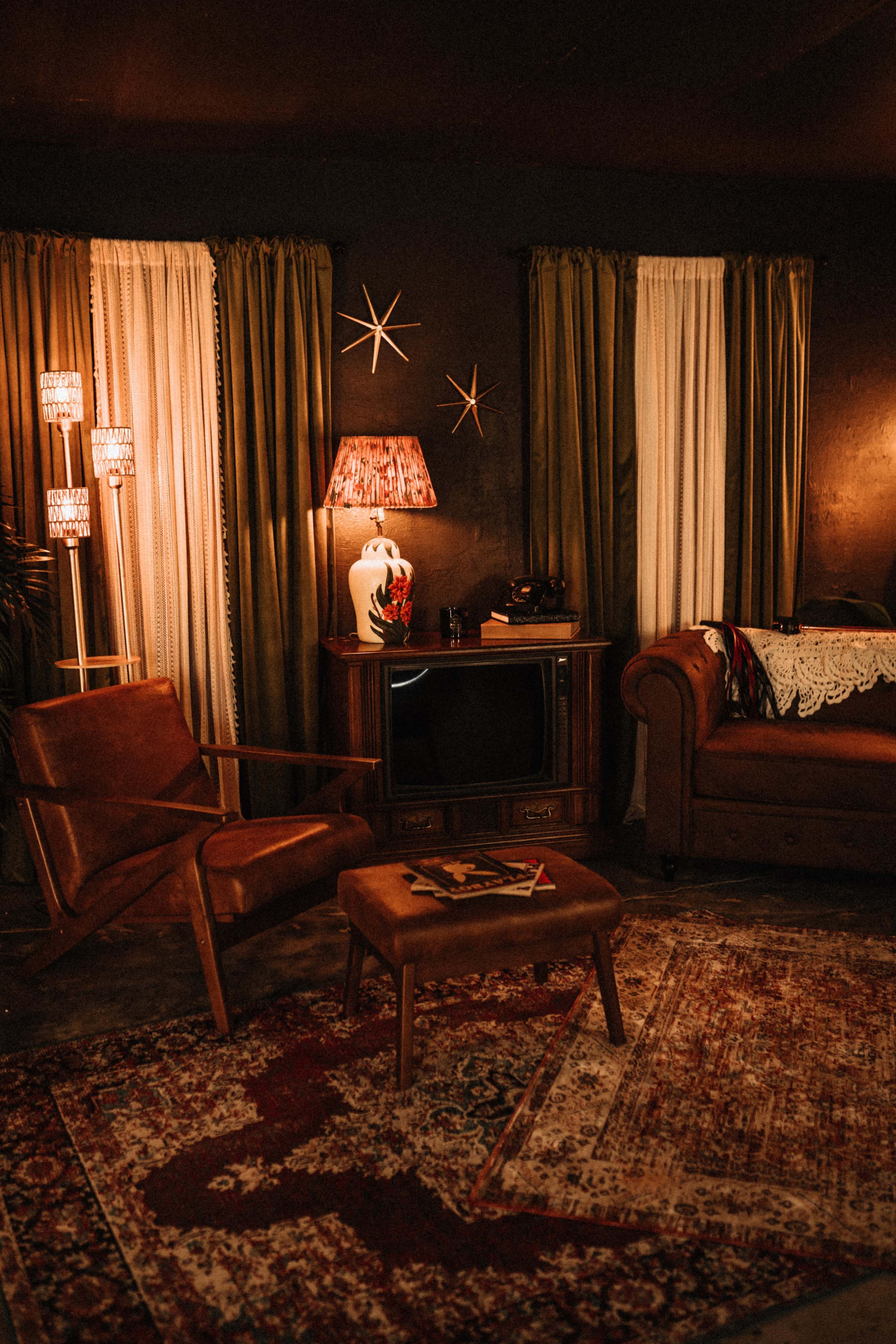 The image shows a cozy living room scene featuring two brown leather chairs, a vintage television, and a lamp, with textured curtains and a decorative area rug.