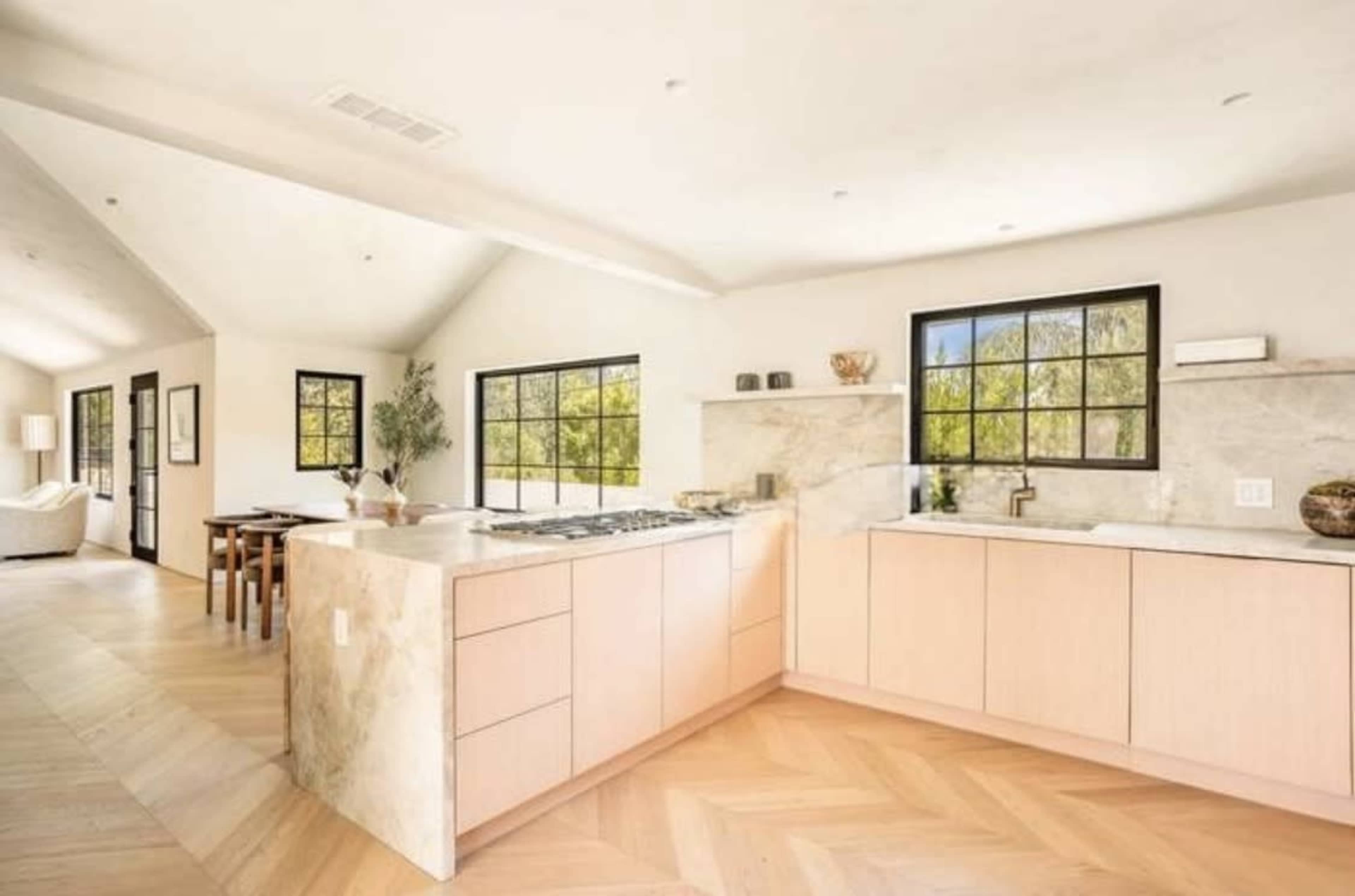 The image shows a modern kitchen with light-colored cabinetry, a large island, and natural wood flooring, open to a dining area and living space.