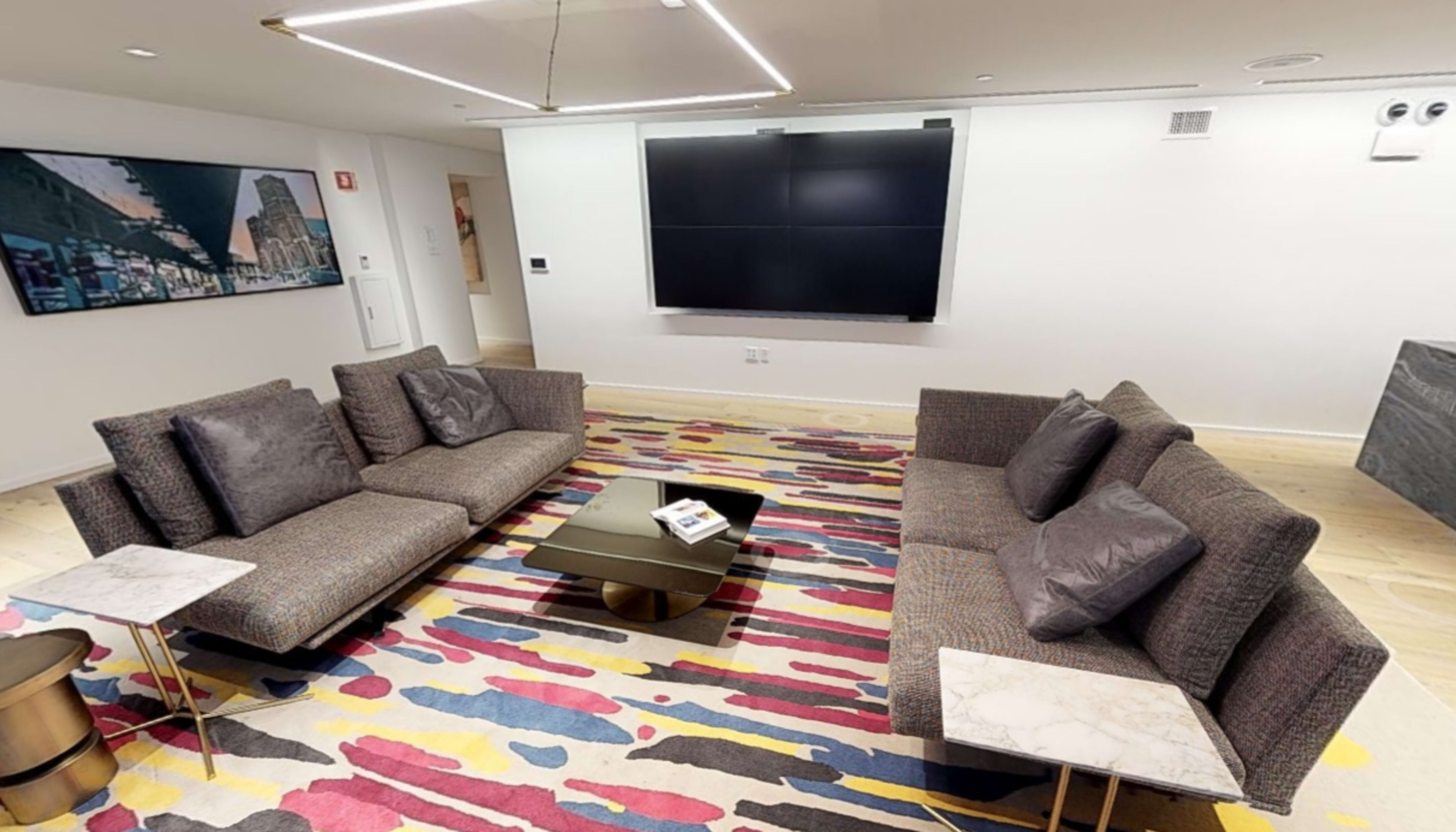 A modern living room with two gray sofas arranged around a black coffee table on a colorful rug, and a wall-mounted TV in the background.
