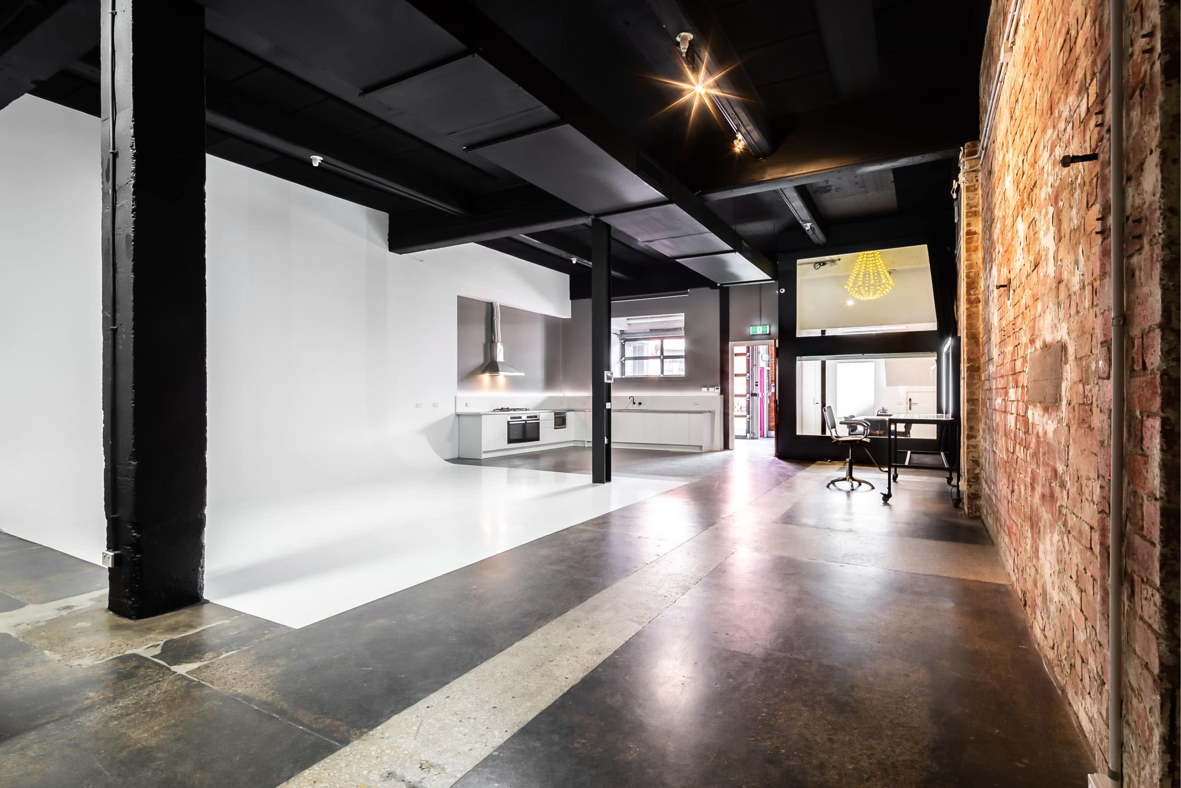 A modern, open-plan space features a kitchen area with sleek appliances, exposed brick walls, and a combination of polished concrete and white flooring.
