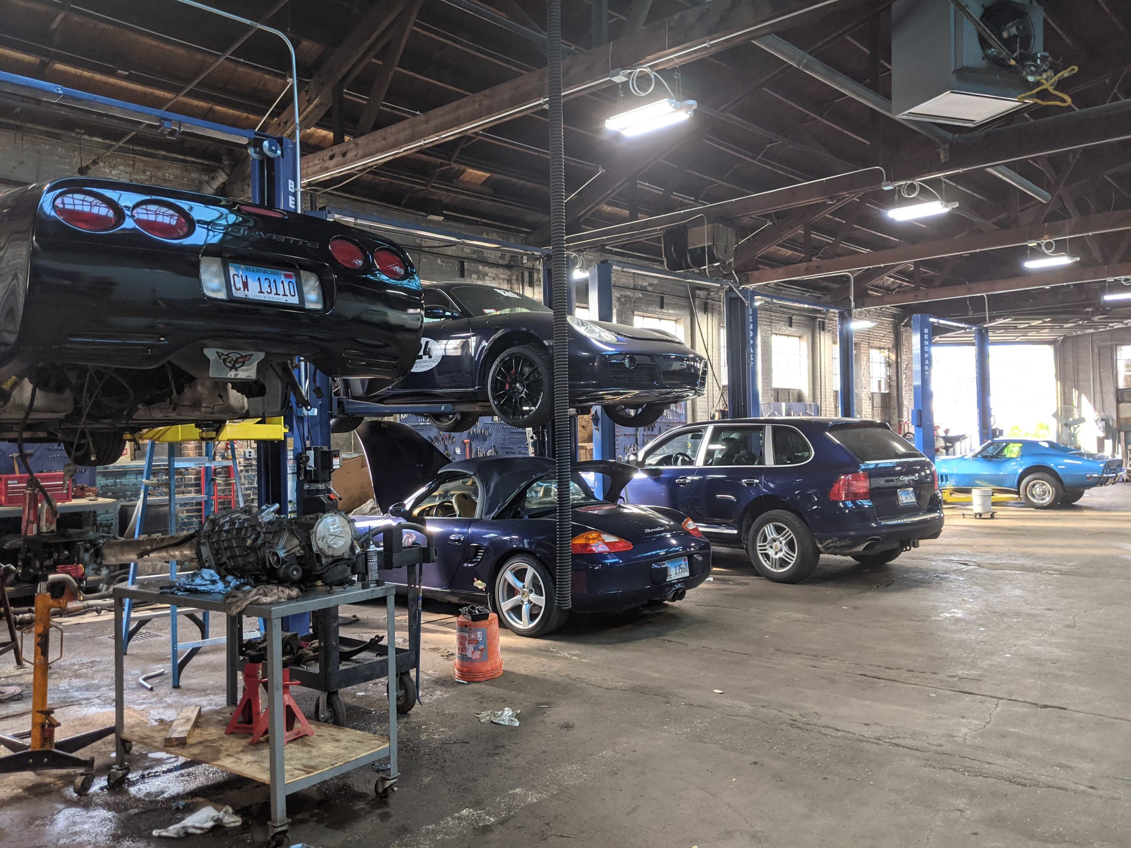 A car repair shop with several vehicles in various stages of maintenance, including lifted cars, a workbench, and tools scattered throughout the space.