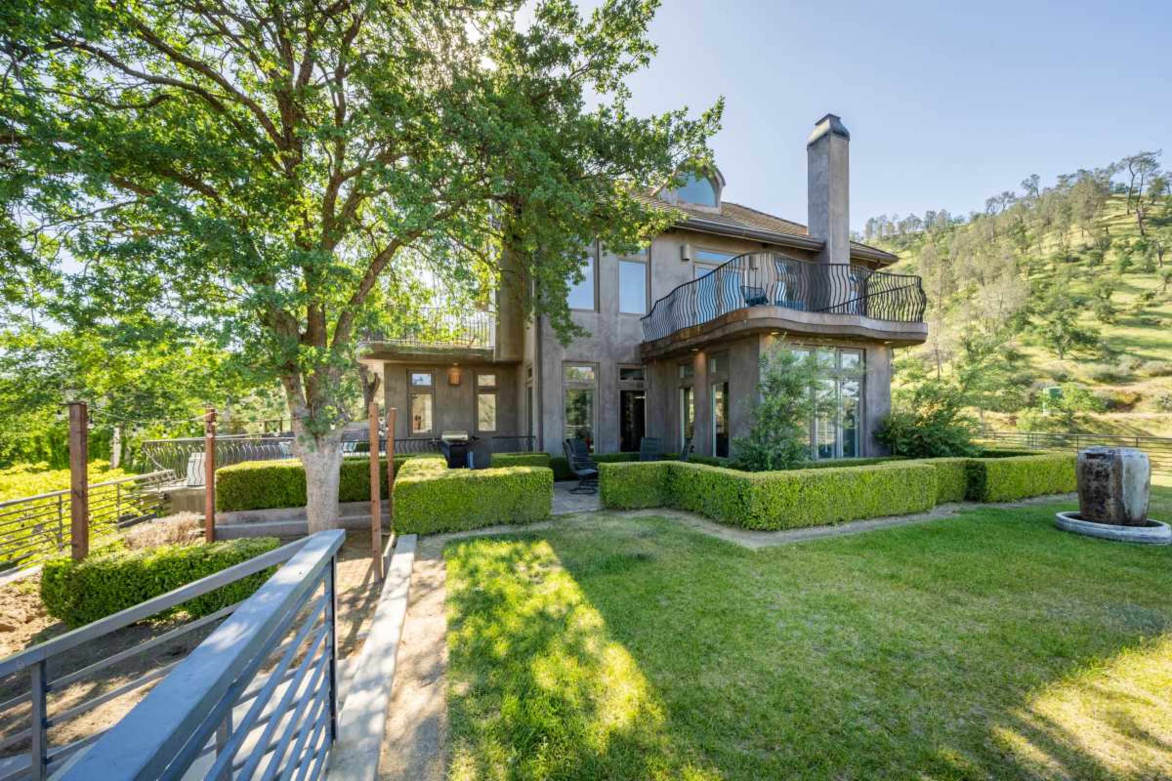 The image shows a two-story house surrounded by manicured hedges and trees, with a sloped hillside in the background.