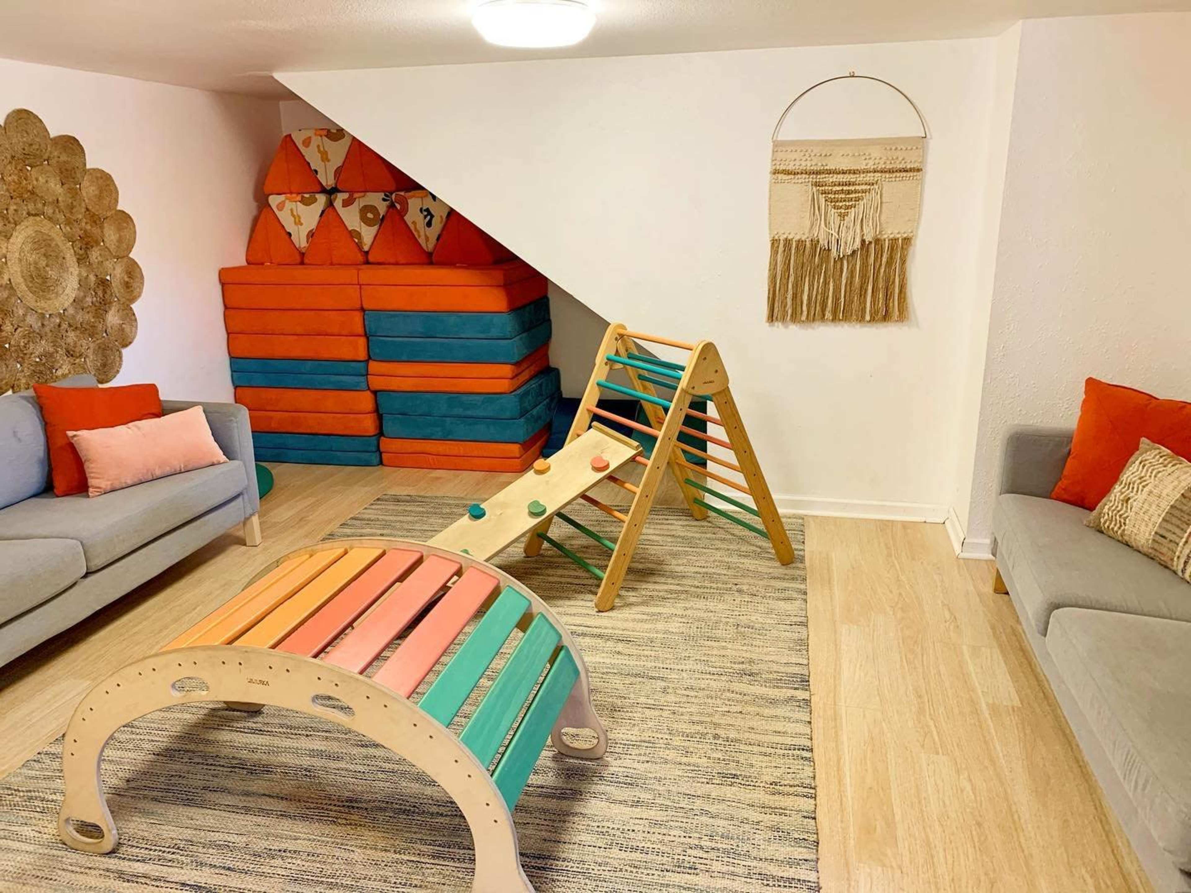 A playroom featuring colorful climbing and balancing equipment, stacked cushions, and two sofas arranged on a light wooden floor.