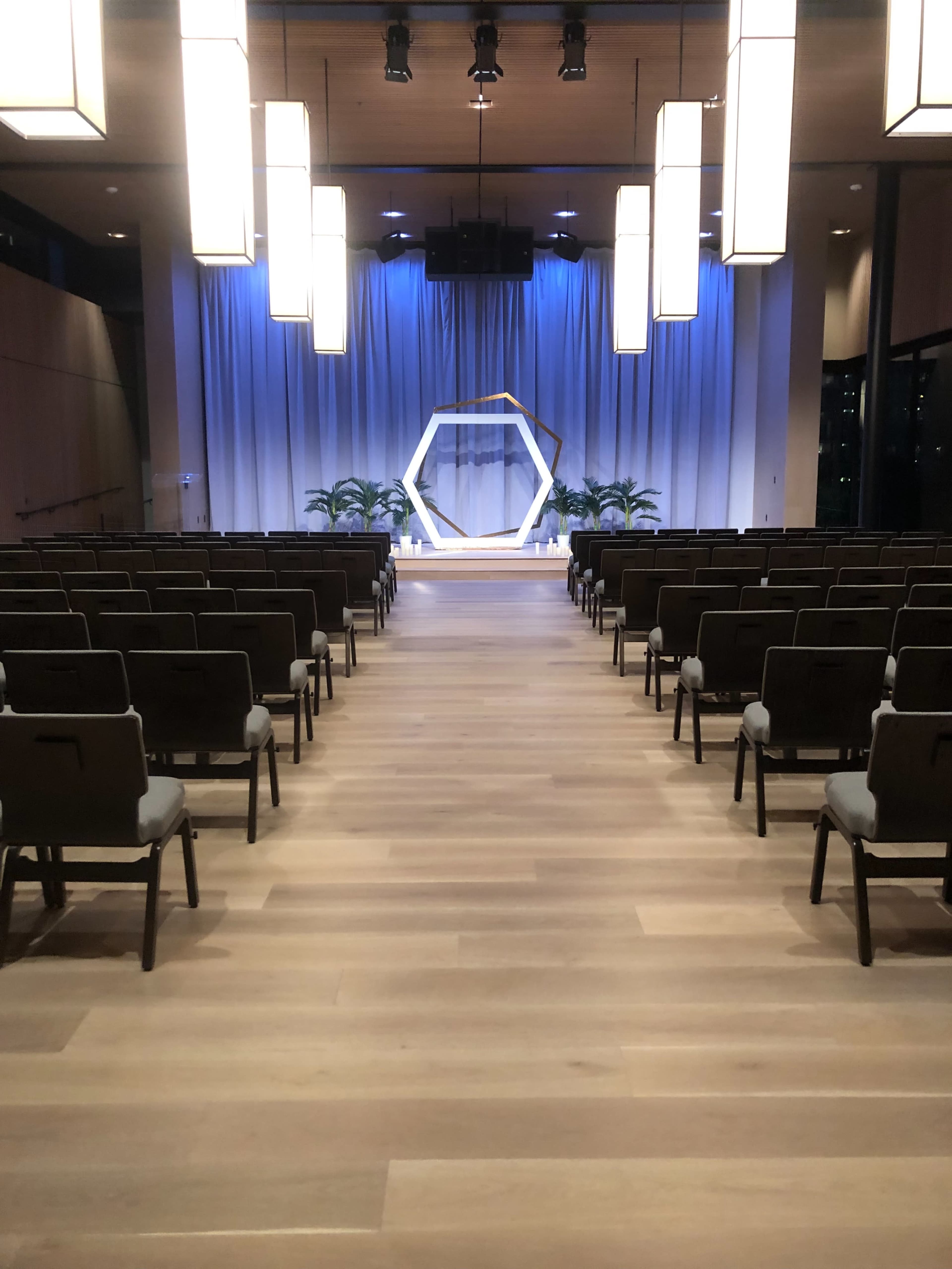 A modern auditorium with rows of chairs facing a stage featuring a hexagonal backdrop and decorative plants.