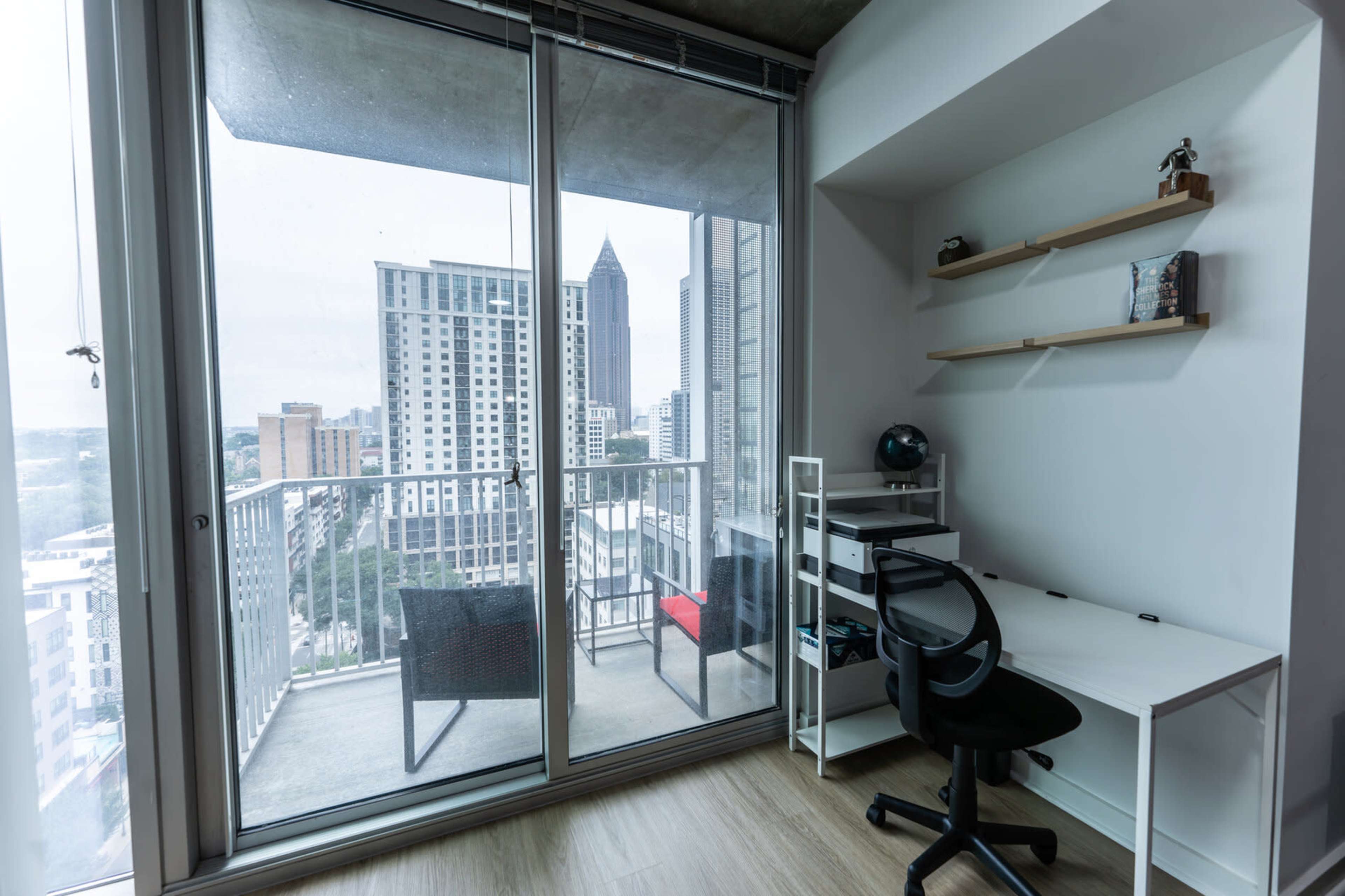 The image shows a modern office space with a desk and chair positioned near a large glass sliding door that opens to a balcony, overlooking a cityscape.