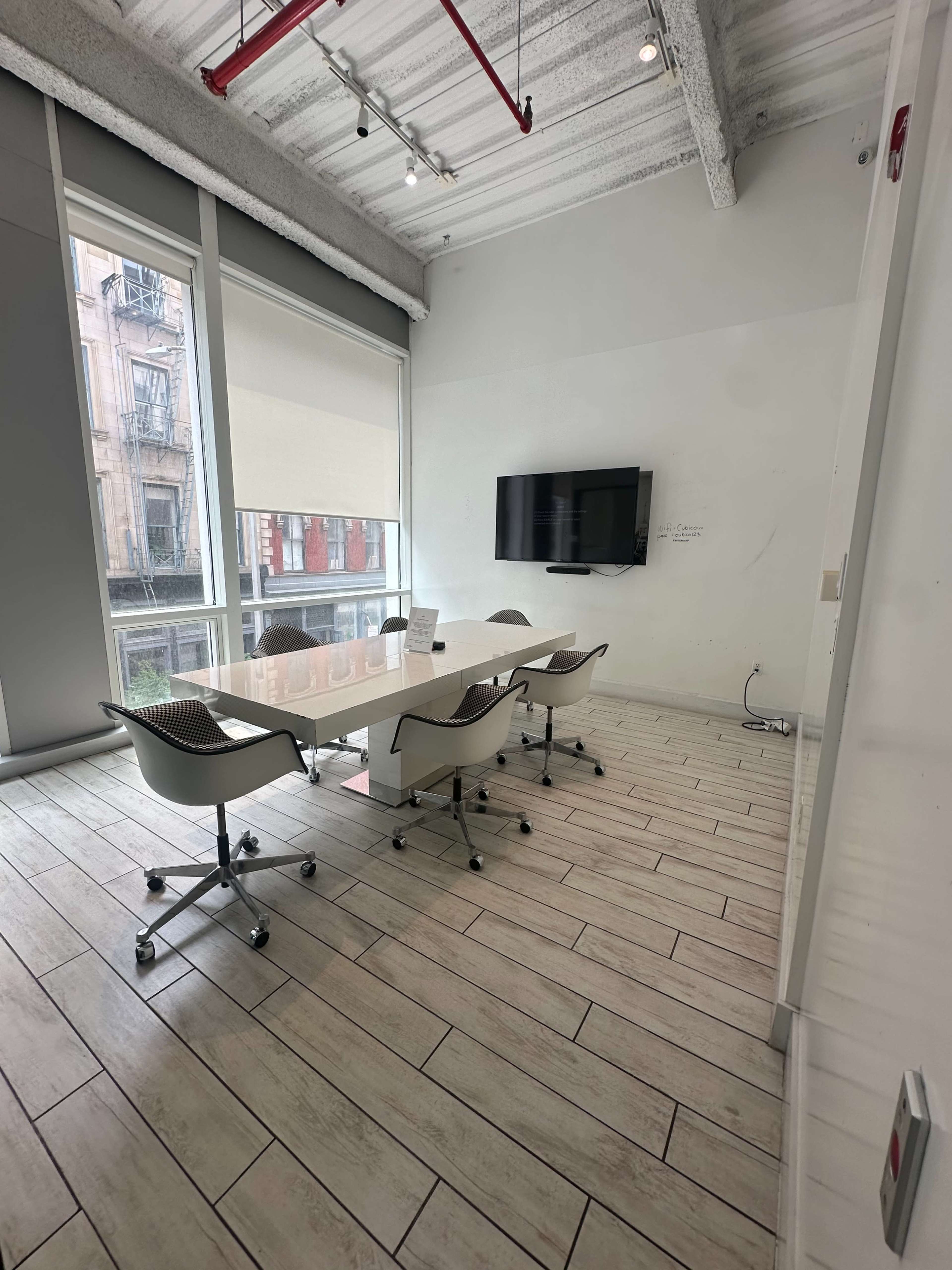 The image shows a modern conference room with a long table, several chairs, a television mounted on the wall, and large windows providing natural light.
