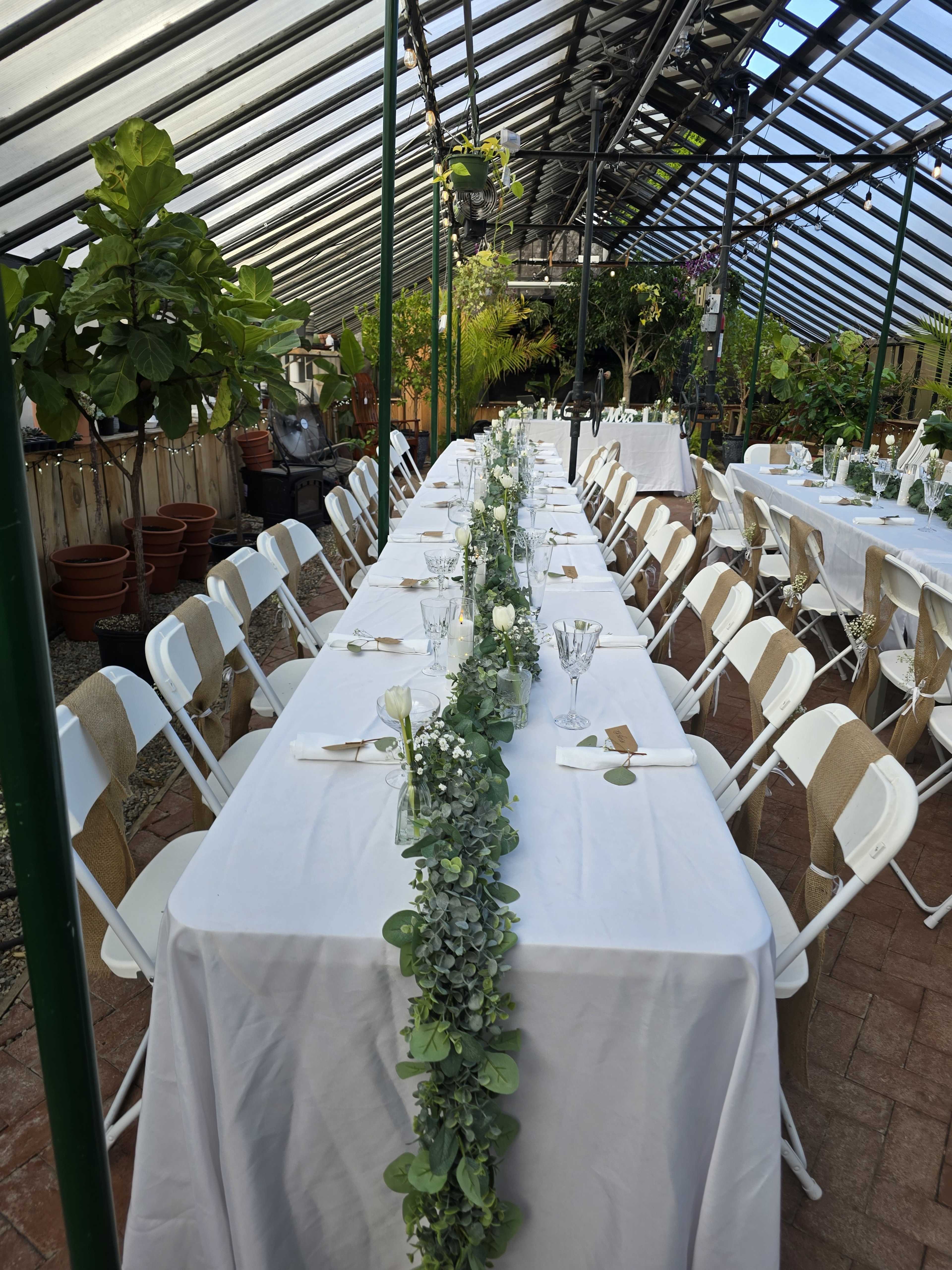 The image shows a long dining table set up in a greenhouse, adorned with a floral runner and surrounded by white chairs.