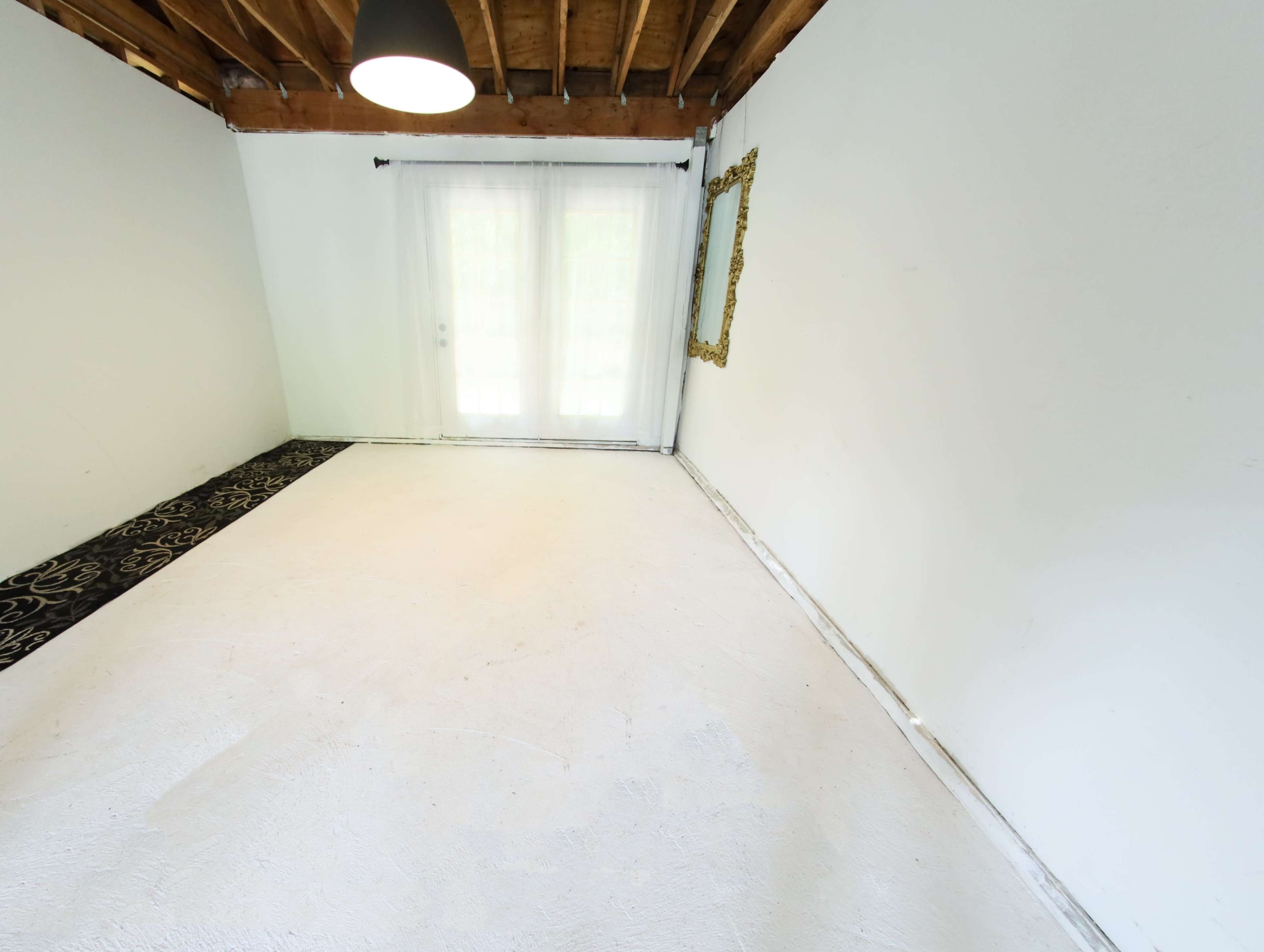 The room features a bare white floor, light walls, a large window with sheer curtains, and a decorative mirror on the wall.