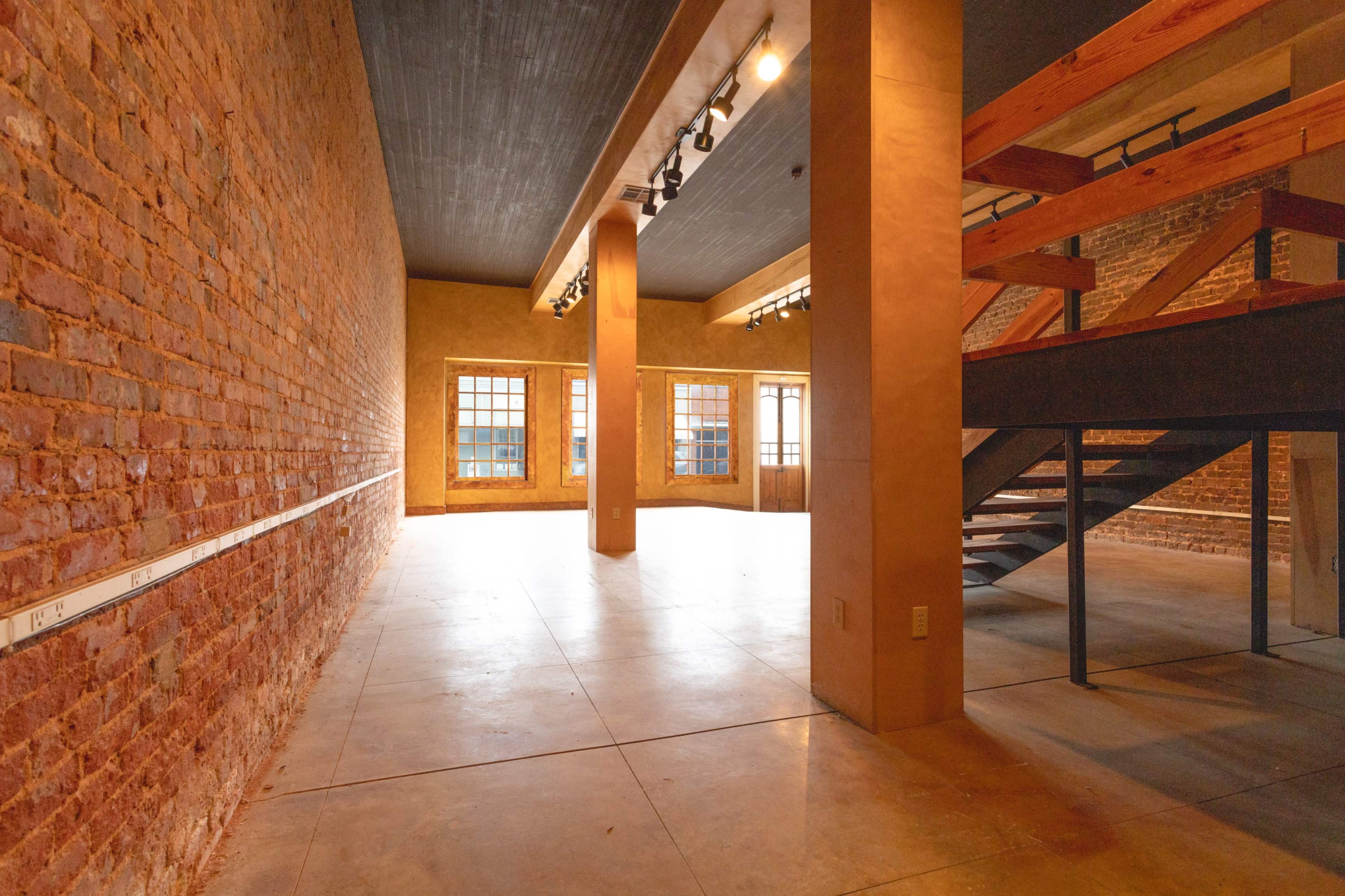 The image shows an empty, spacious interior with exposed brick walls, concrete flooring, and a wooden staircase leading to a loft area.