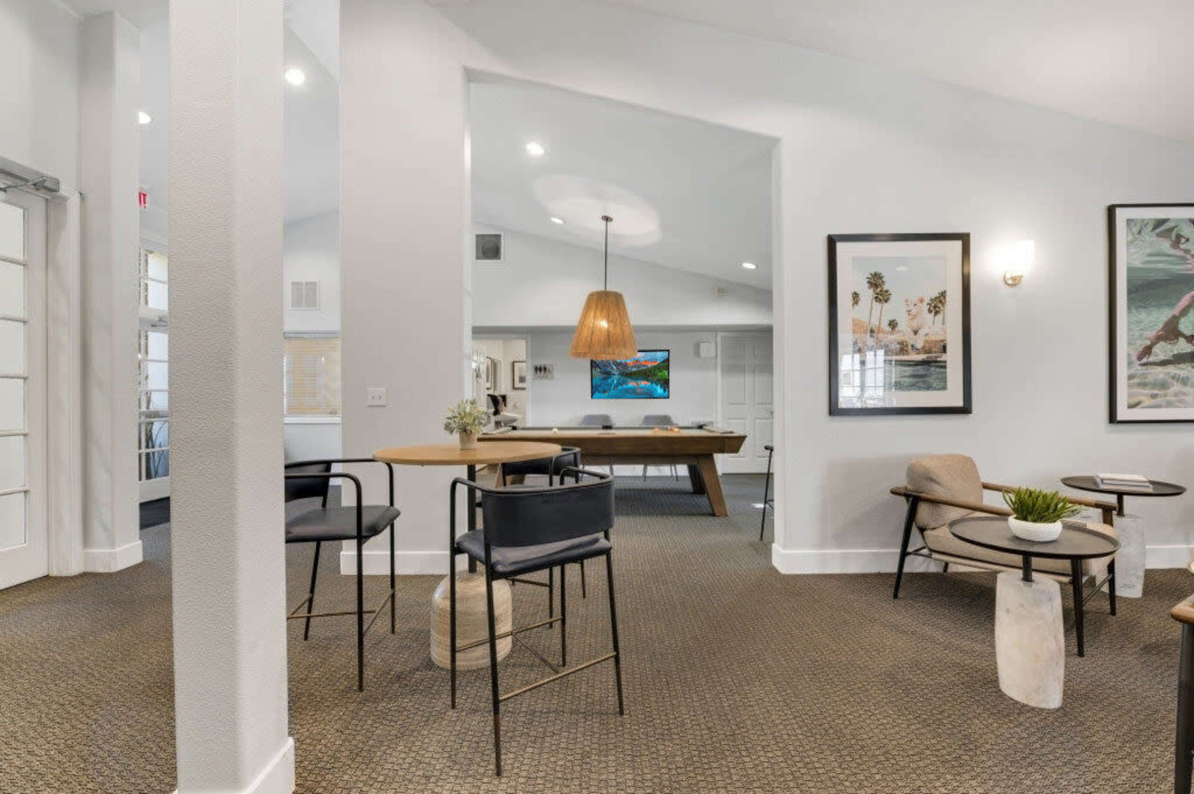 The image shows a modern interior space with a seating area, a wooden dining table, and various decorative artworks on the walls.
