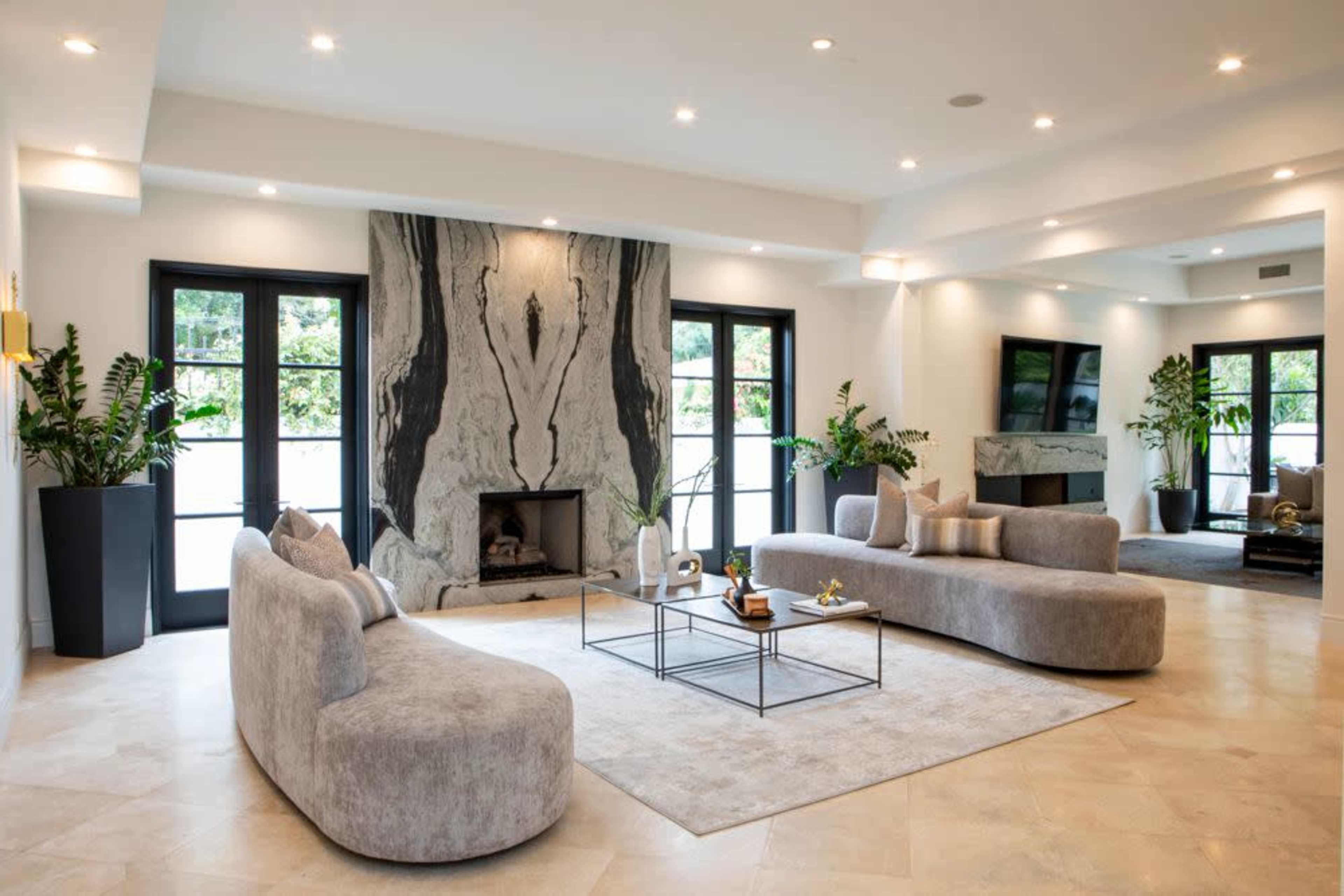 A modern living room features a large, curved gray sofa, a matching loveseat, a black coffee table, a stone fireplace, and large windows surrounded by potted plants.