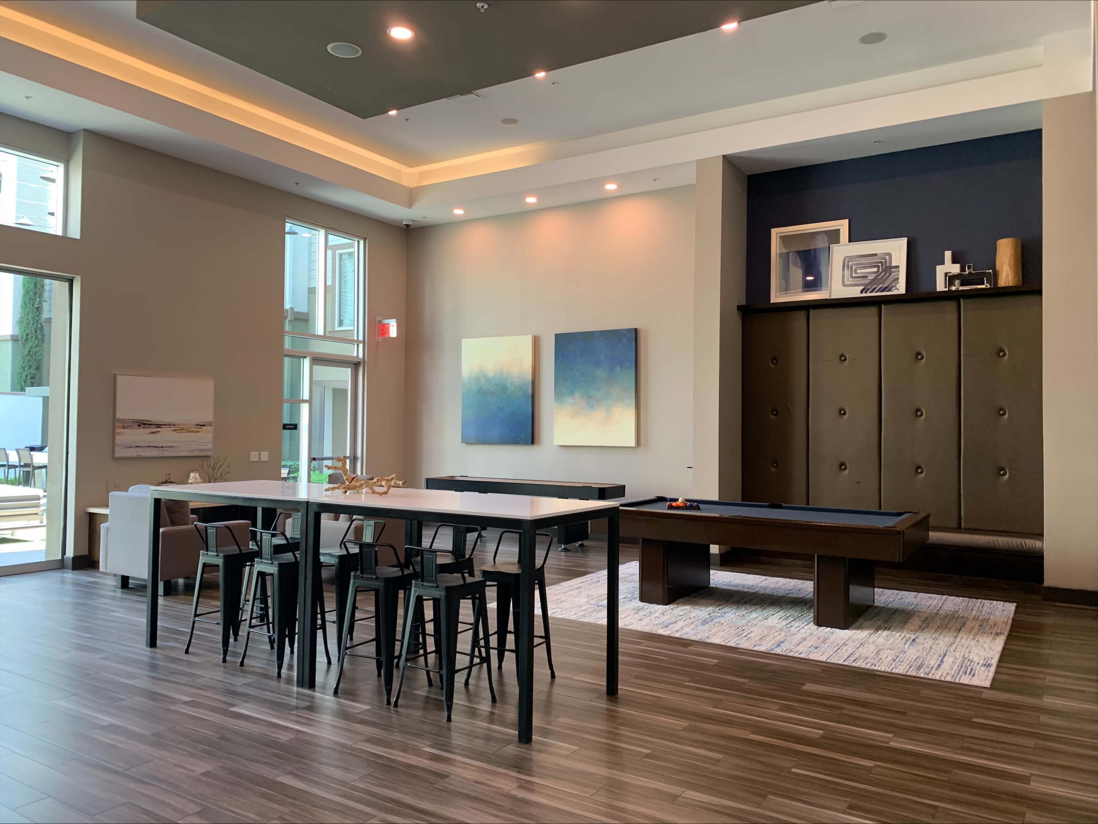 A modern communal lounge area with a dining table, barstool seating, a billiards table, and abstract artwork hanging on the walls.