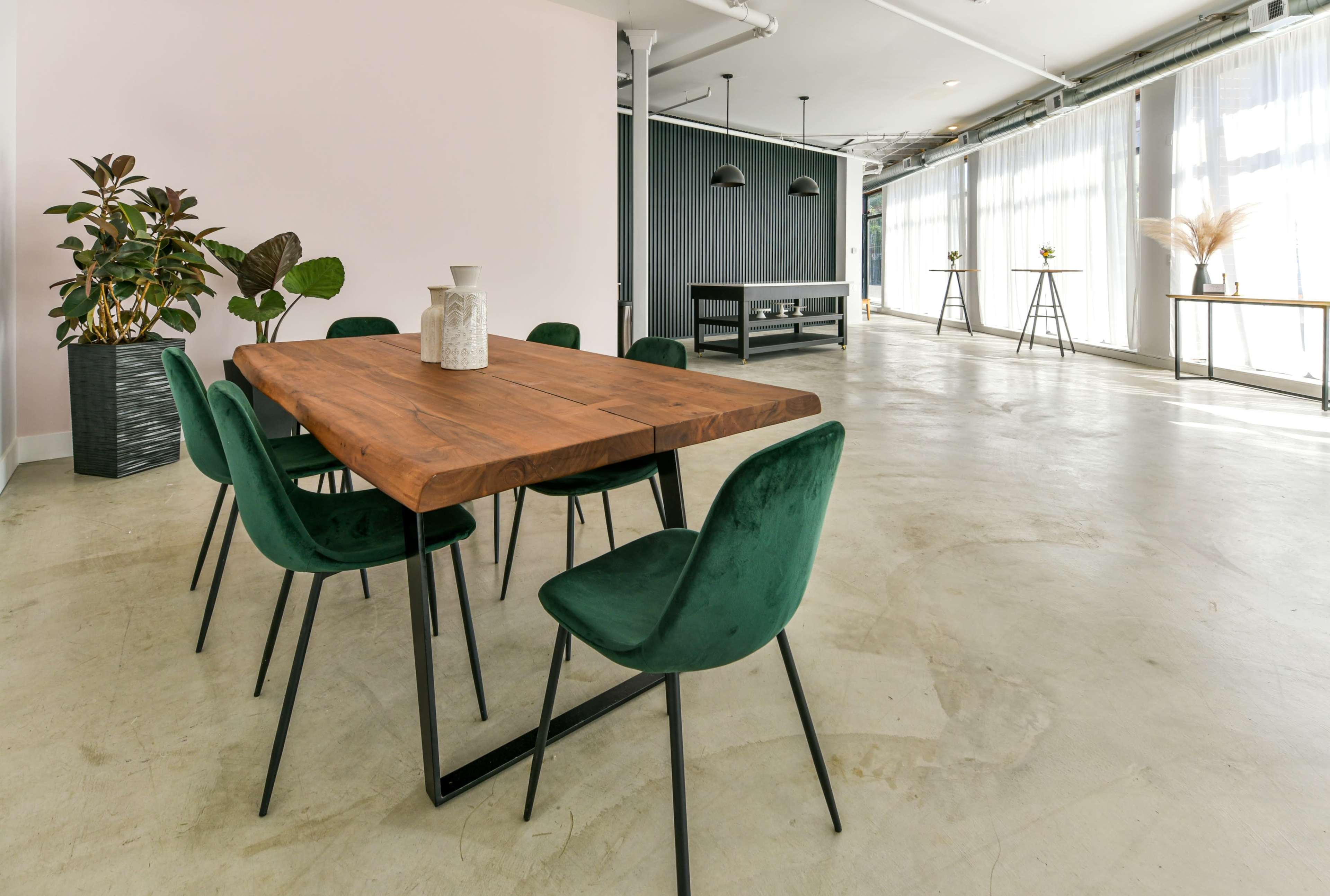 A wooden dining table with green upholstered chairs is positioned in a spacious, brightly lit room featuring large windows and minimalist decor.