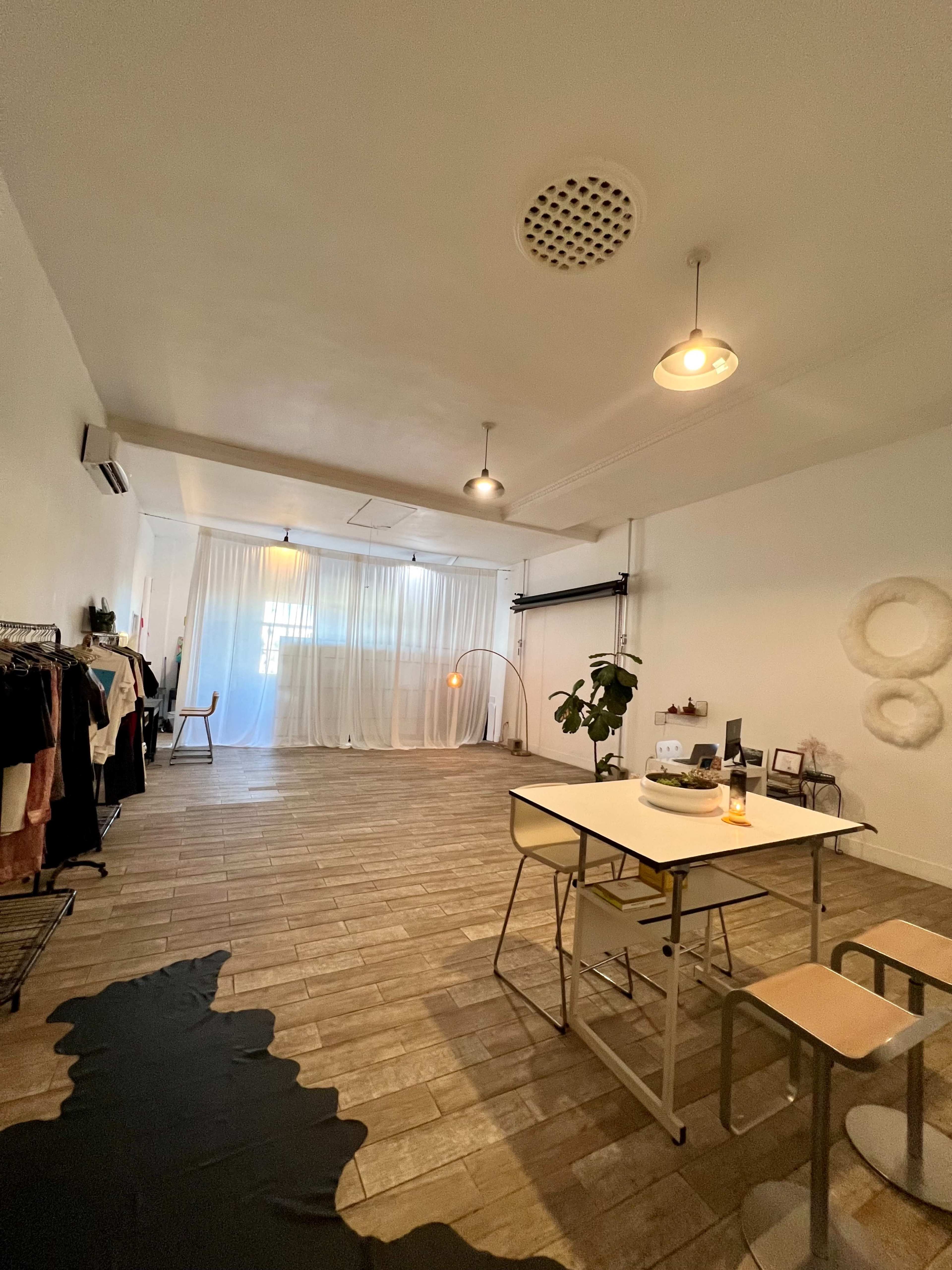 A spacious room with wooden flooring, a dining table with two chairs, and clothing racks along one wall, illuminated by soft lighting and curtains in the background.