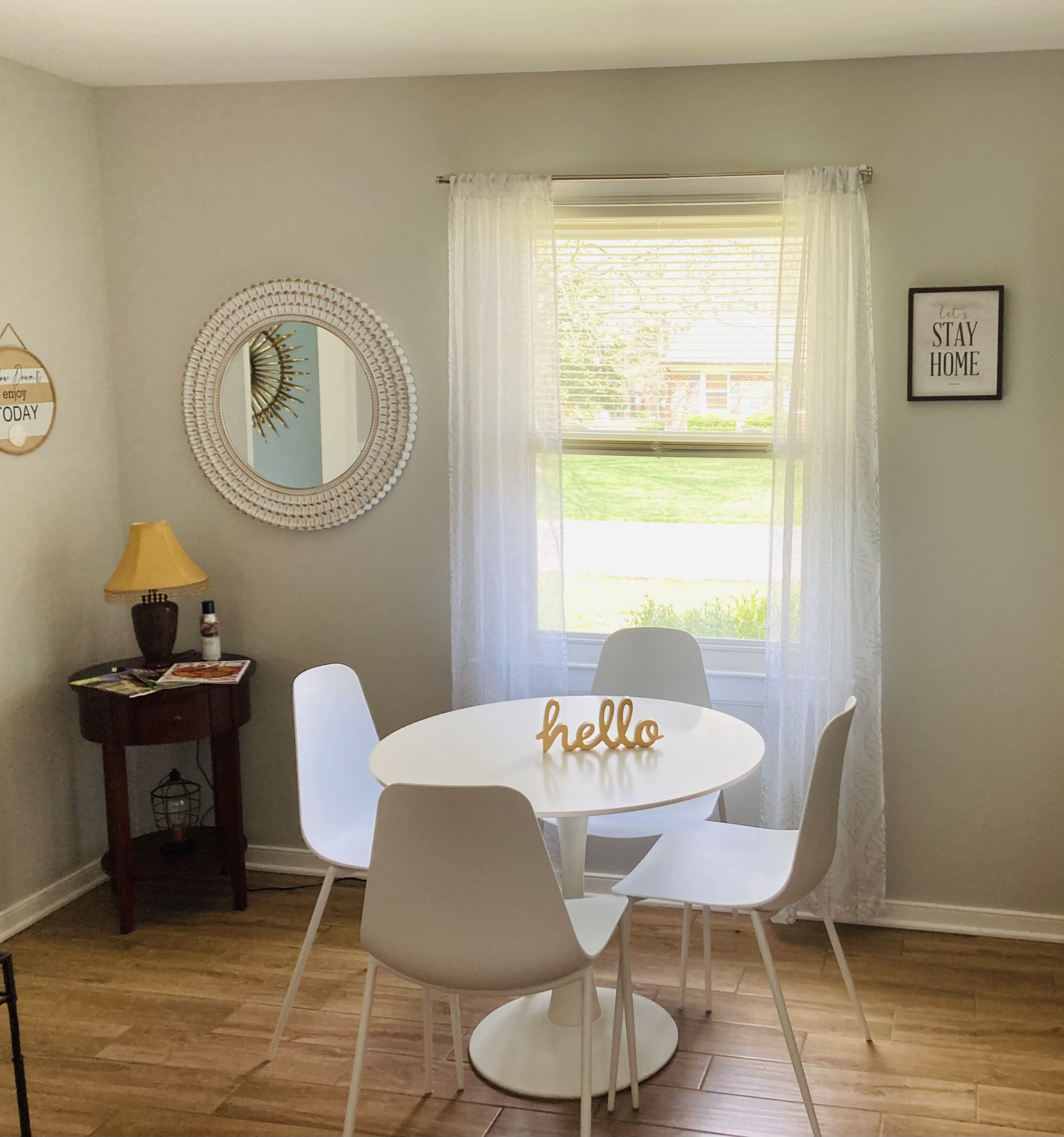 A small dining area features a round white table surrounded by four white chairs, with a lamp and decorative items nearby, and a window letting in natural light.