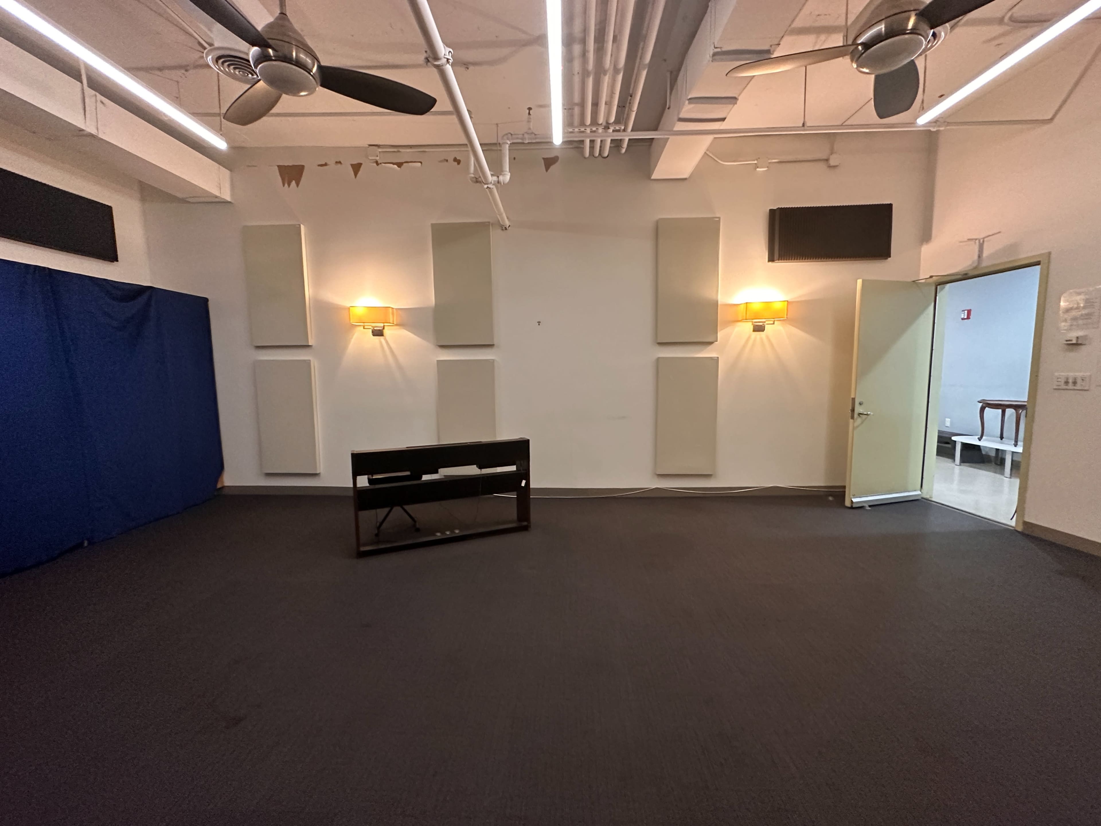 An empty room features a dark floor, wall-mounted lights, acoustic panels, and a door leading to another space.