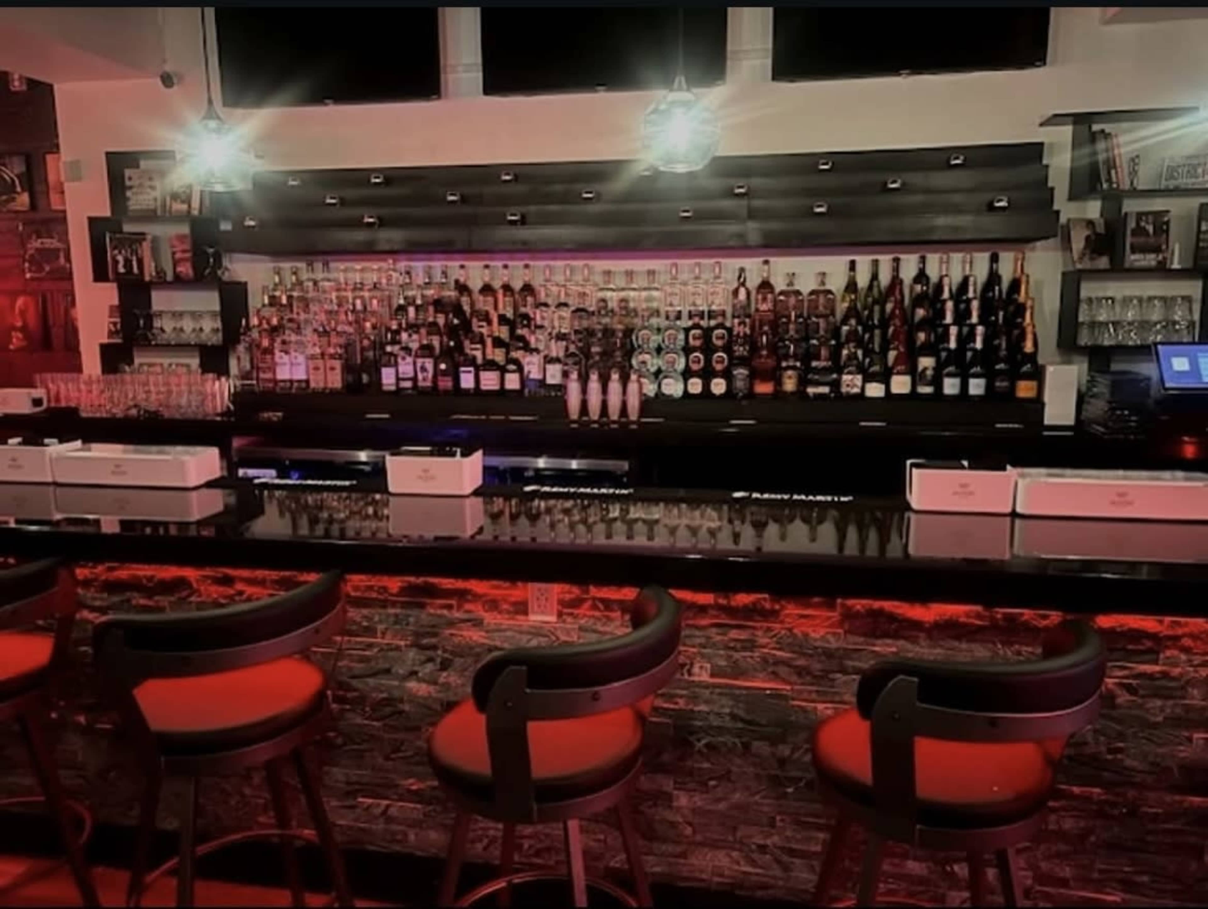 The image shows a well-lit bar with a long countertop, featuring an extensive array of liquor bottles displayed on shelves behind it.
