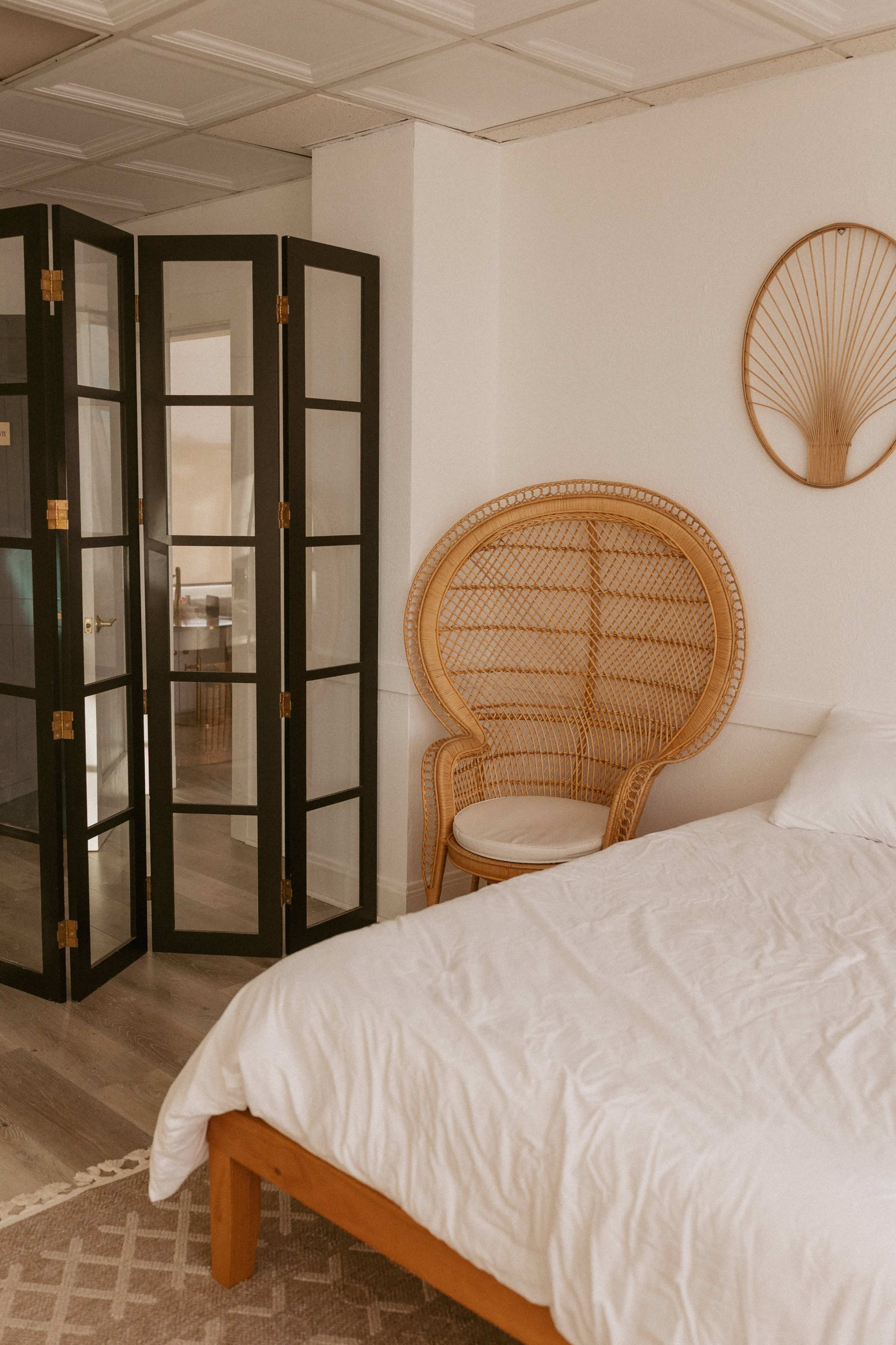 The image shows a cozy bedroom featuring a bed with a white comforter, a rattan chair, and a folding screen in the background.